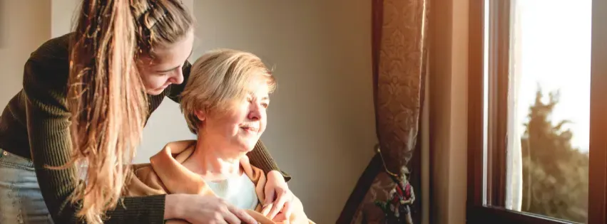 A young person with long hair puts their hands on the shoulders of an older person, who looks out a window.