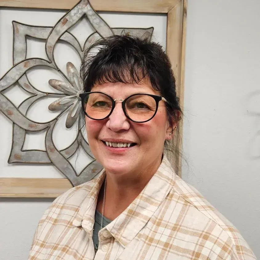 Woman wearing glasses, smiling, plaid shirt, in front of a decorative metal wall art piece.