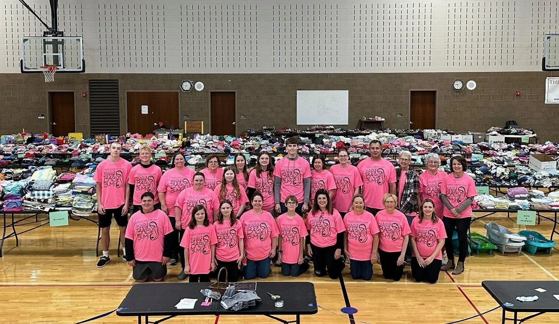 Group of people in pink shirts pose in a gymnasium in front of tables filled with donations.