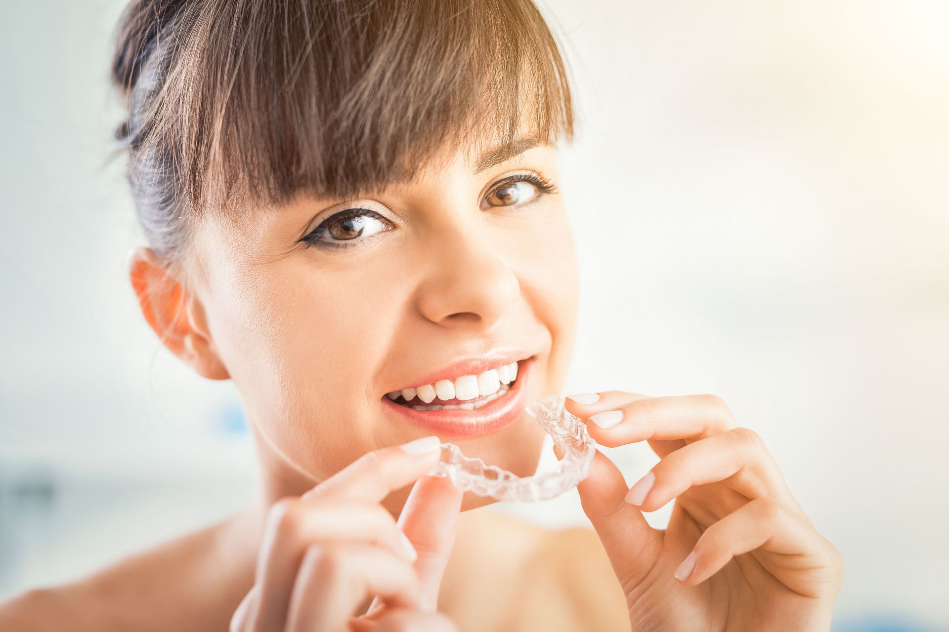 Woman holding clear teeth aligner, smiling, close-up. Bright background.