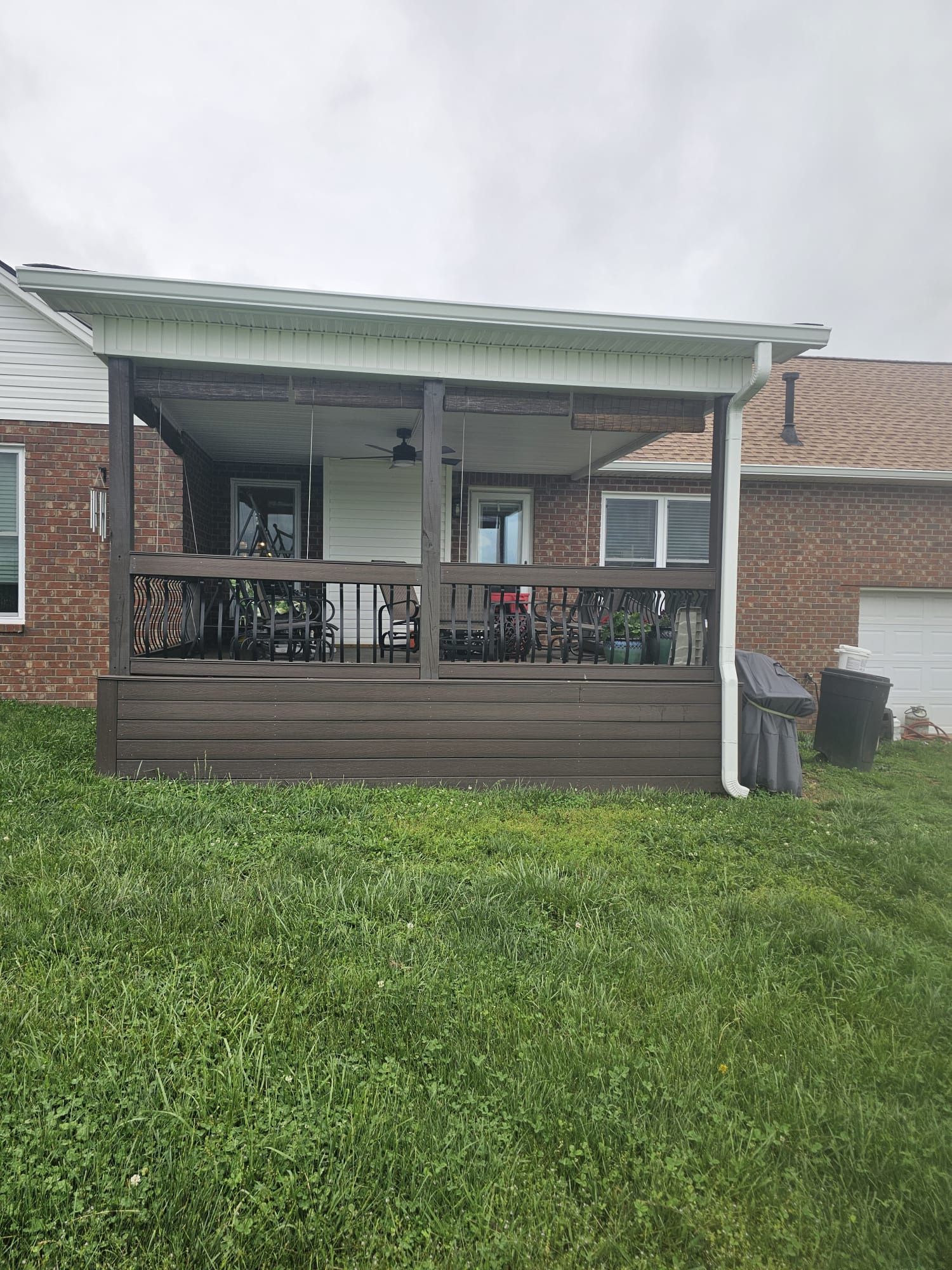A backyard wooden deck with a covered roof, brown railings, and furniture, attached to a brick house.