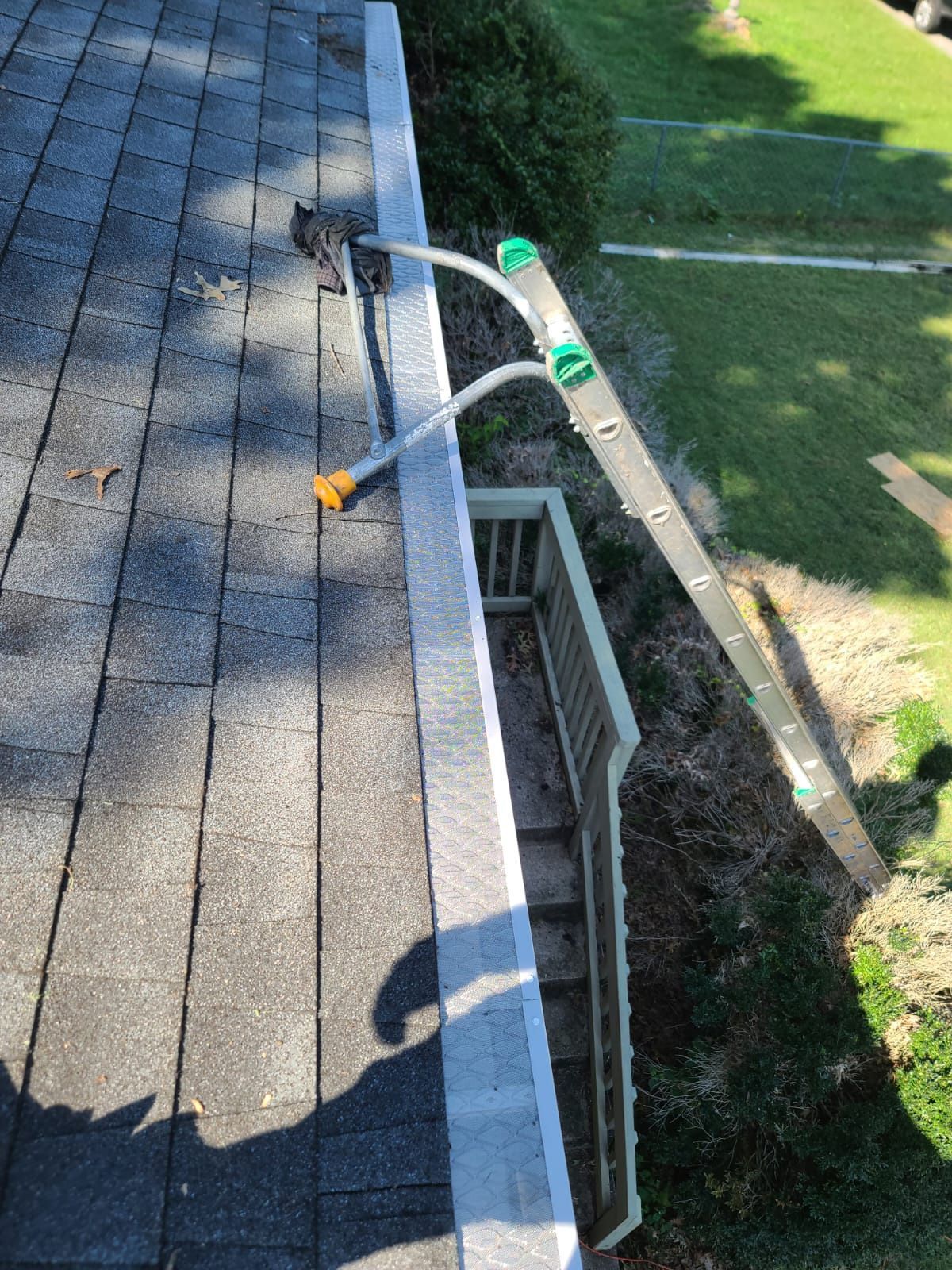 A ladder stabilizer rests on the edge of a roof near a gutter, with the ladder extended down toward the ground below.