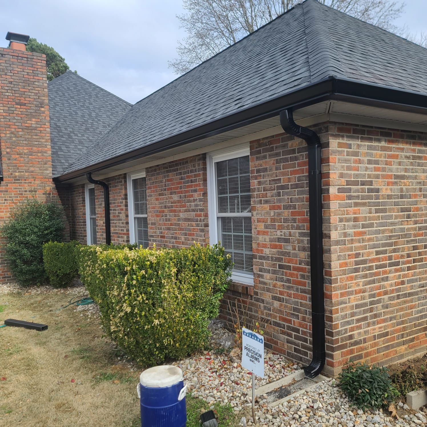 A side view of a brick house with a new black roof and black gutters, featuring two windows and landscaping shrubs.