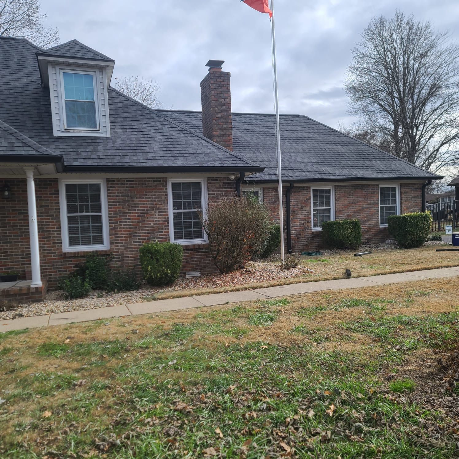 A single-story brick house with a dark shingled roof, a dormer window, a brick chimney, and a tall flagpole on the lawn.
