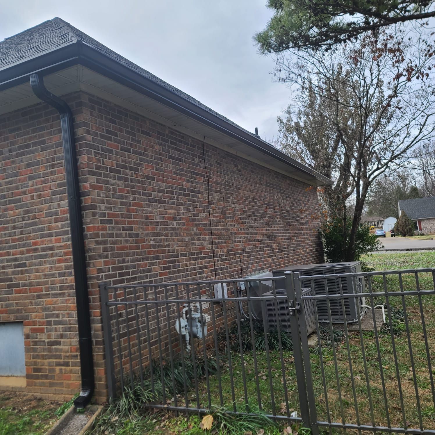 Side view of a brick house exterior showing a vertical wall crack, black downspout, and two HVAC units behind a fence.