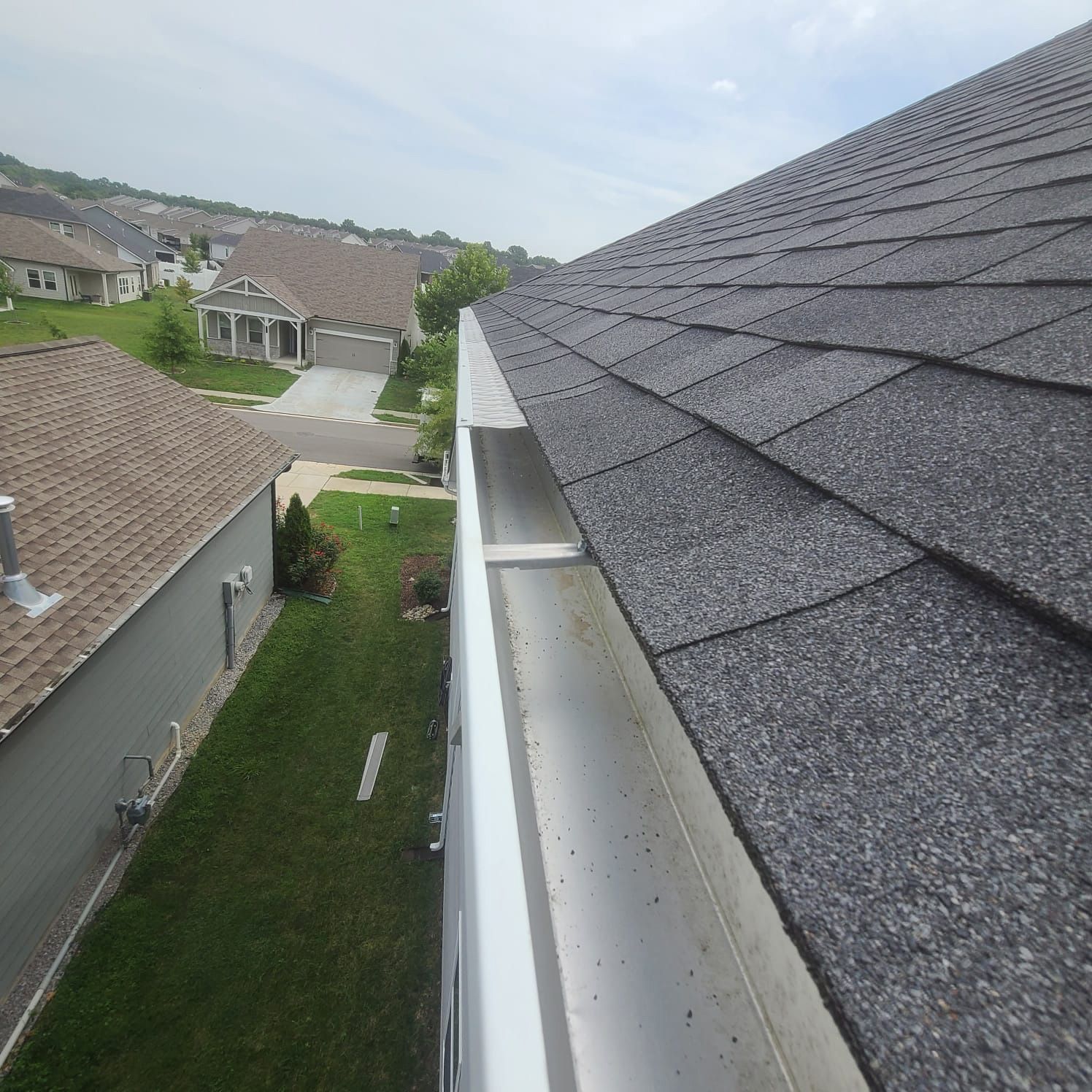 A high-angle view along a clean metal rain gutter running along the edge of a shingled roof above a grassy neighborhood.