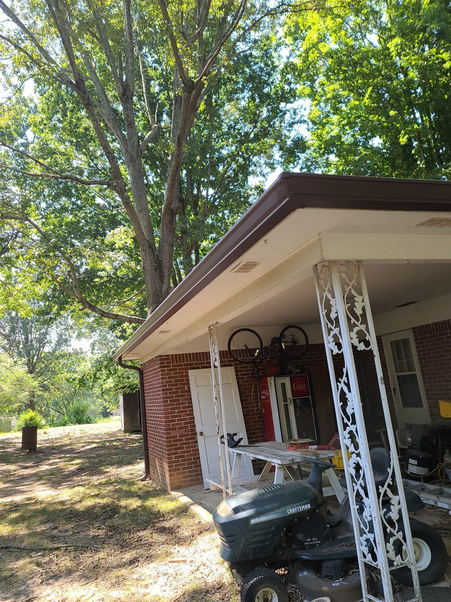 A covered brick patio area featuring white ornamental metal columns, a riding lawnmower, and large trees in the background.