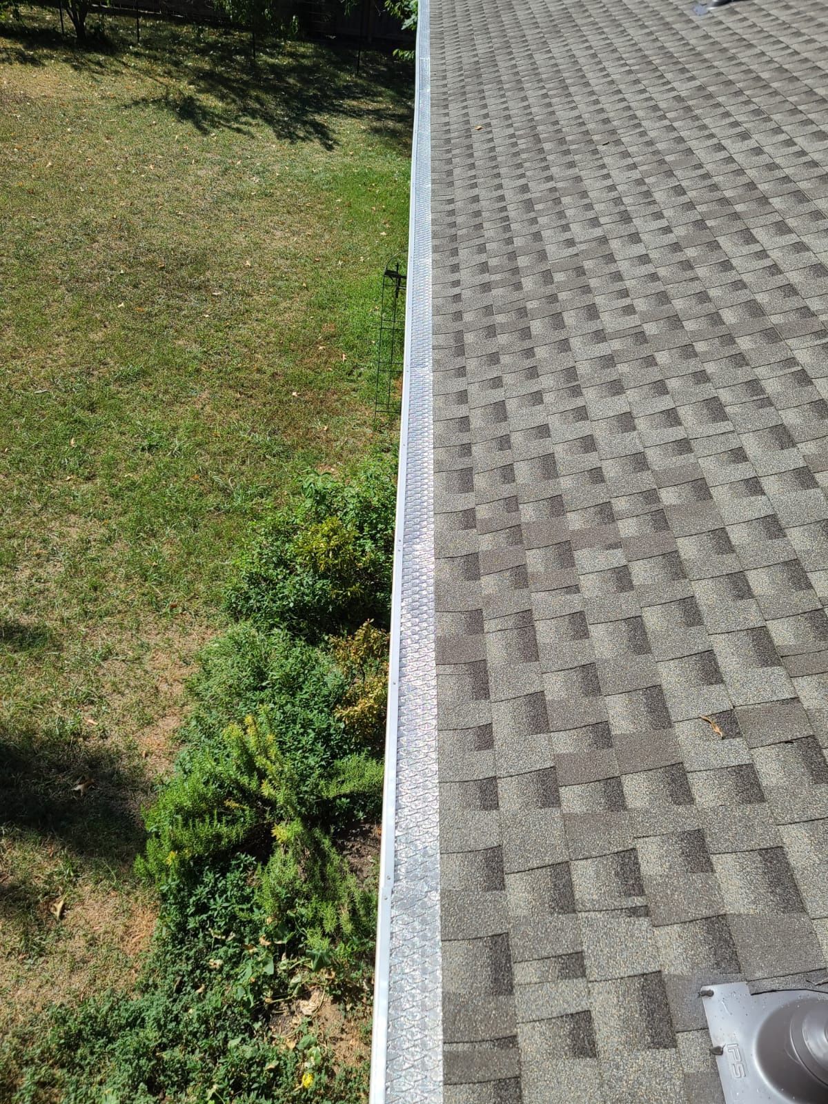 A high-angle view of a gray asphalt shingle roof edge adjacent to a green lawn with a line of small shrubs.