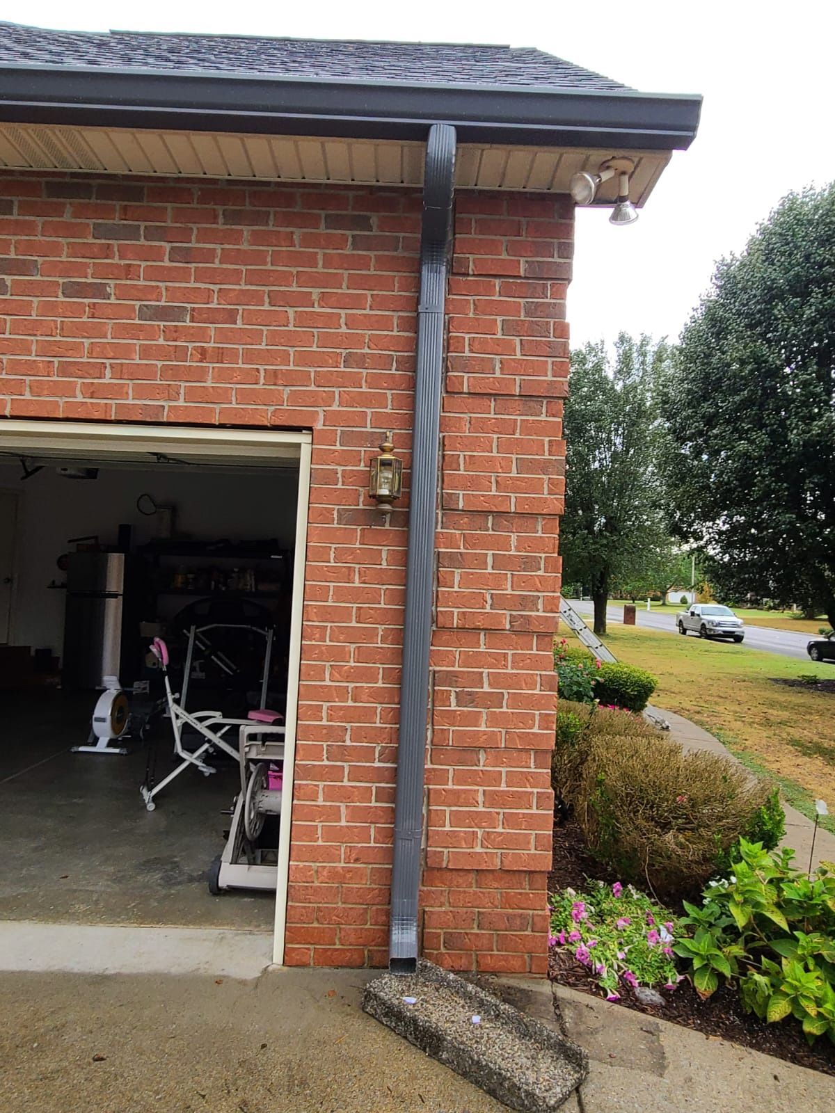 A dark rectangular downspout attached to the brick exterior of a garage, resting on a concrete splash block.