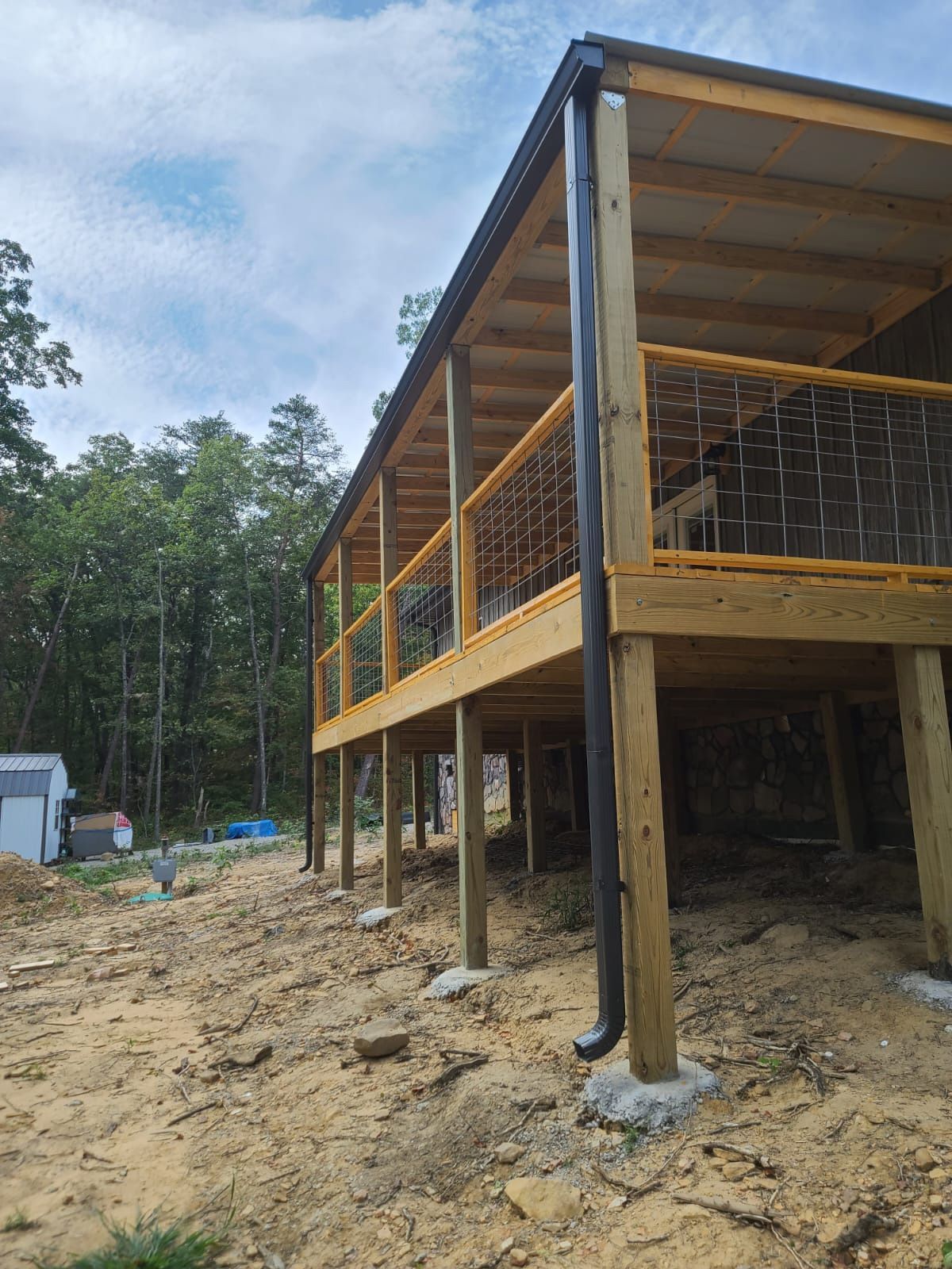 A low-angle view of a wooden deck under construction with metal mesh railings, attached to a house in a wooded area.