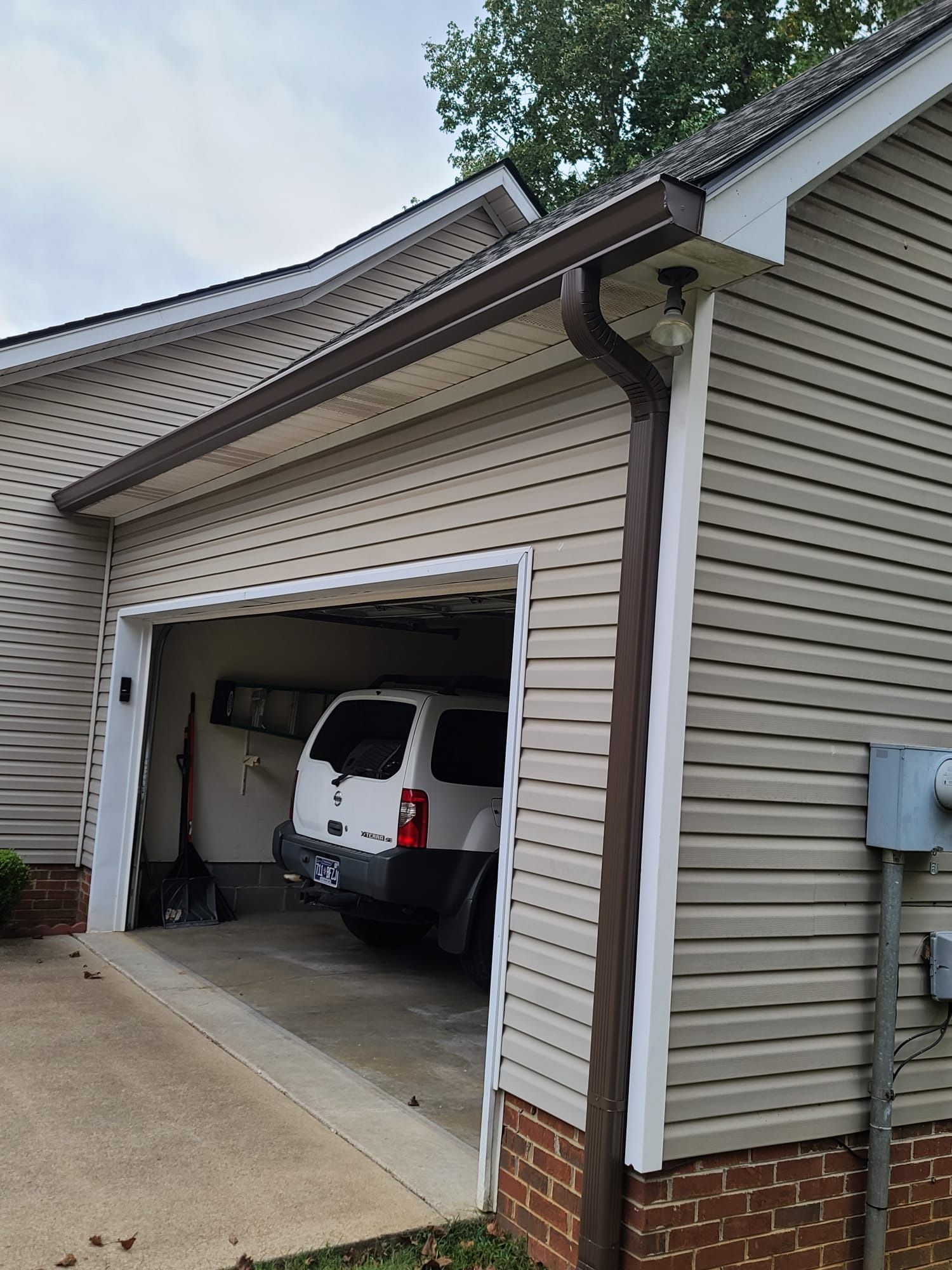 A tan, vinyl-sided garage with a brown gutter system and an open door showing a white SUV parked inside.