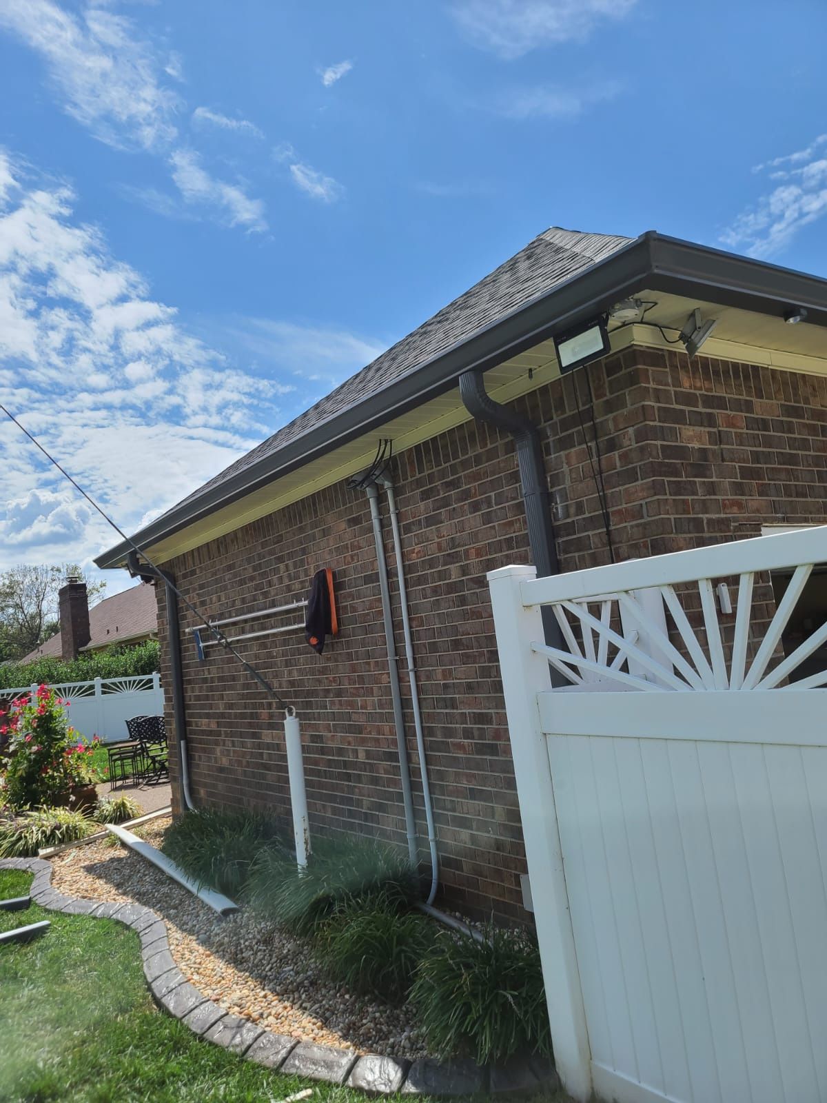 A brick house corner with a gutter downspout, an extended ladder leaning against the wall, and a white fence nearby.