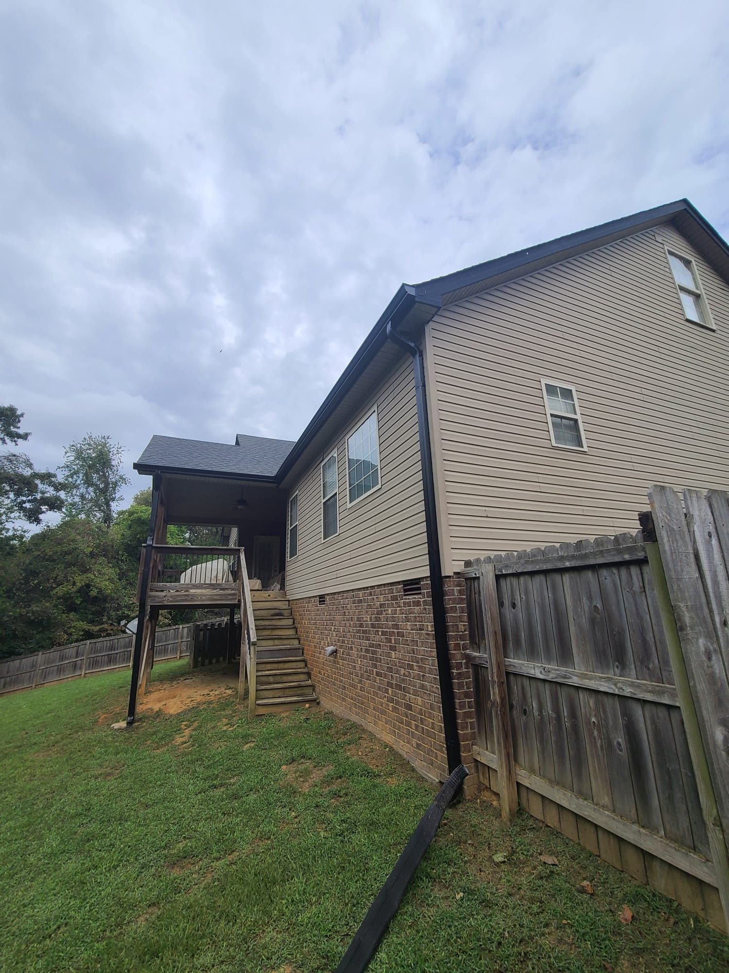 A side view of a tan, two-story house with a brick foundation, a wooden deck with stairs, and a black downspout.