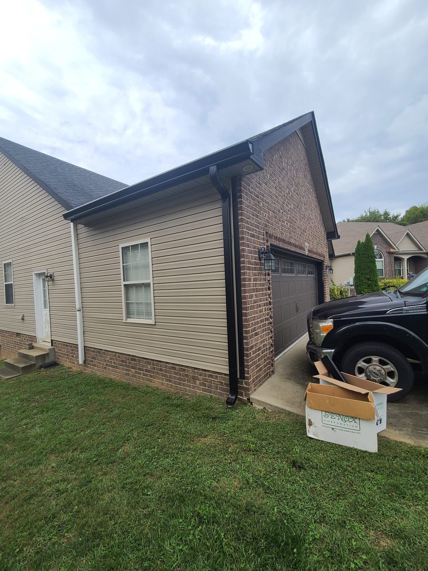 Exterior view of a residential garage with beige siding and brick accents, alongside a black truck and cardboard boxes.
