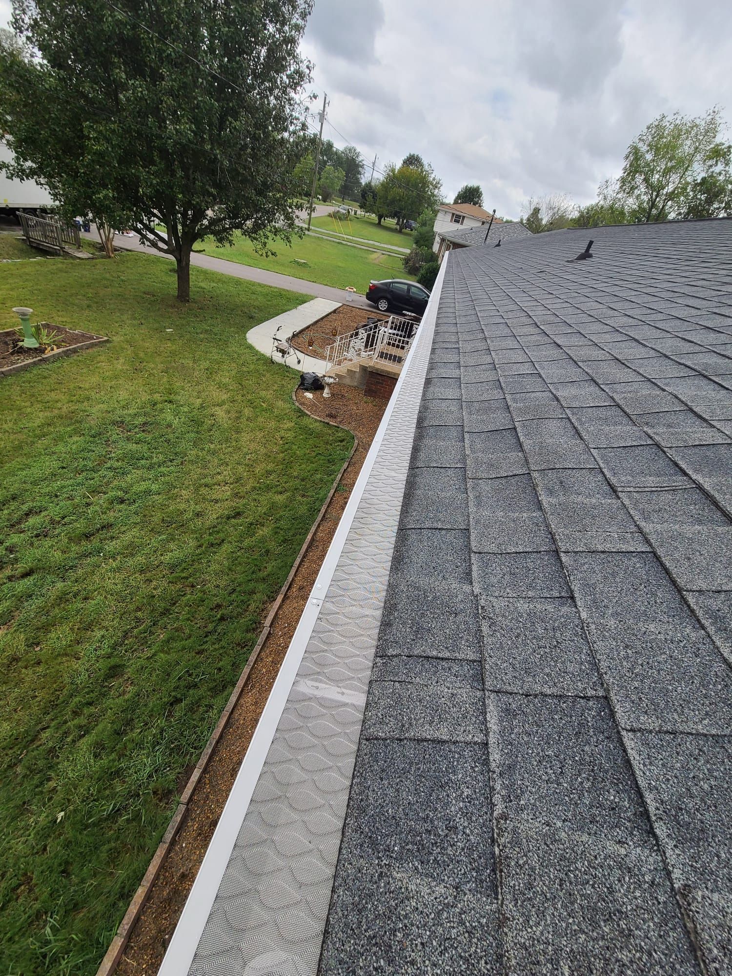 A high-angle view of a gray asphalt shingle roof with white gutter guards installed along the edge, overlooking a yard.