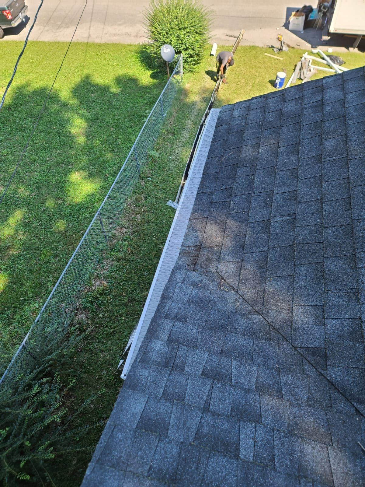An overhead view of a dark shingled roof edge with white gutter guards, beside a grassy yard with a chain-link fence.