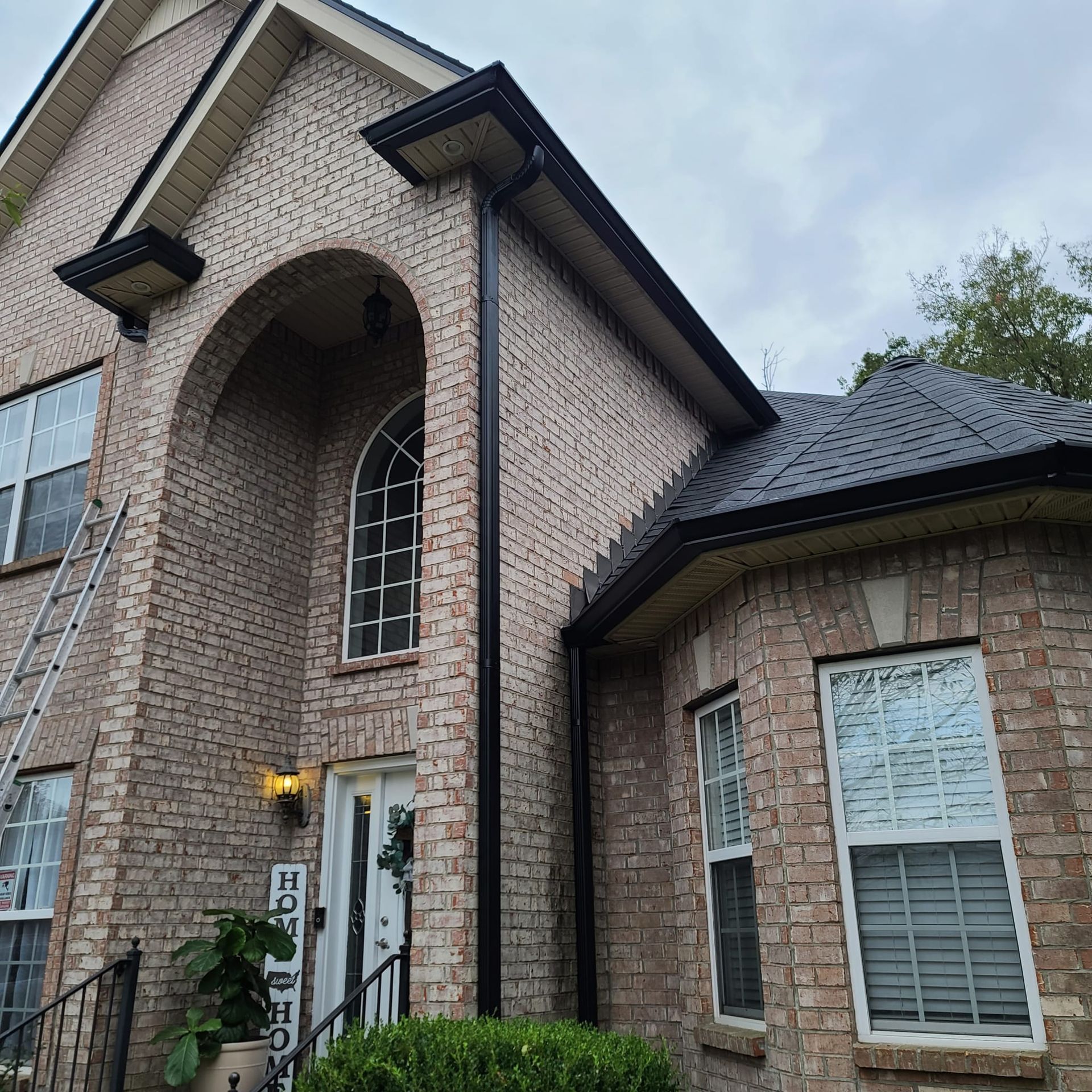 A brick two-story house with a arched entryway, black gutters, and a ladder leaning against the left side.