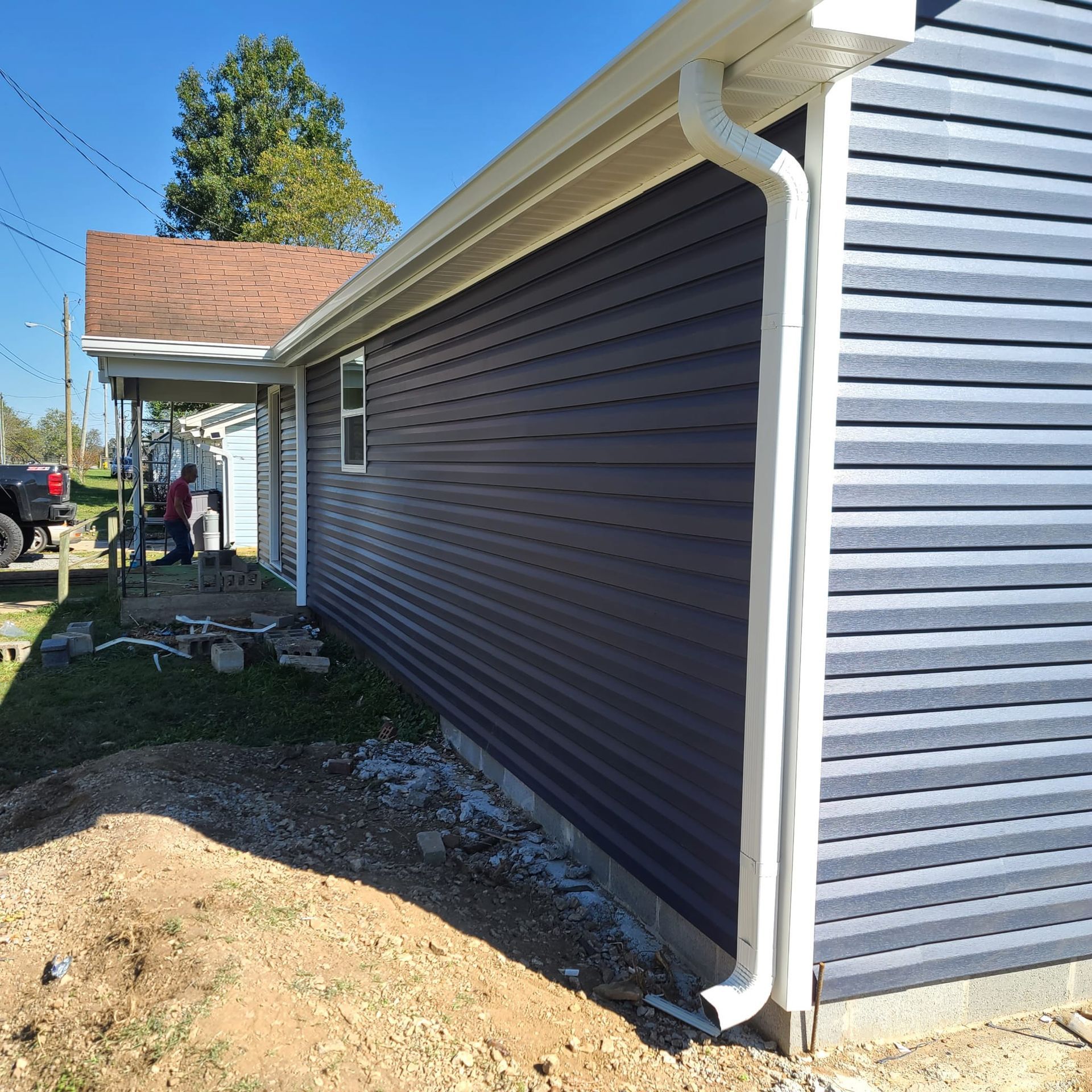A side view of a house with dark gray horizontal siding, a white gutter system, and an unfinished dirt yard.