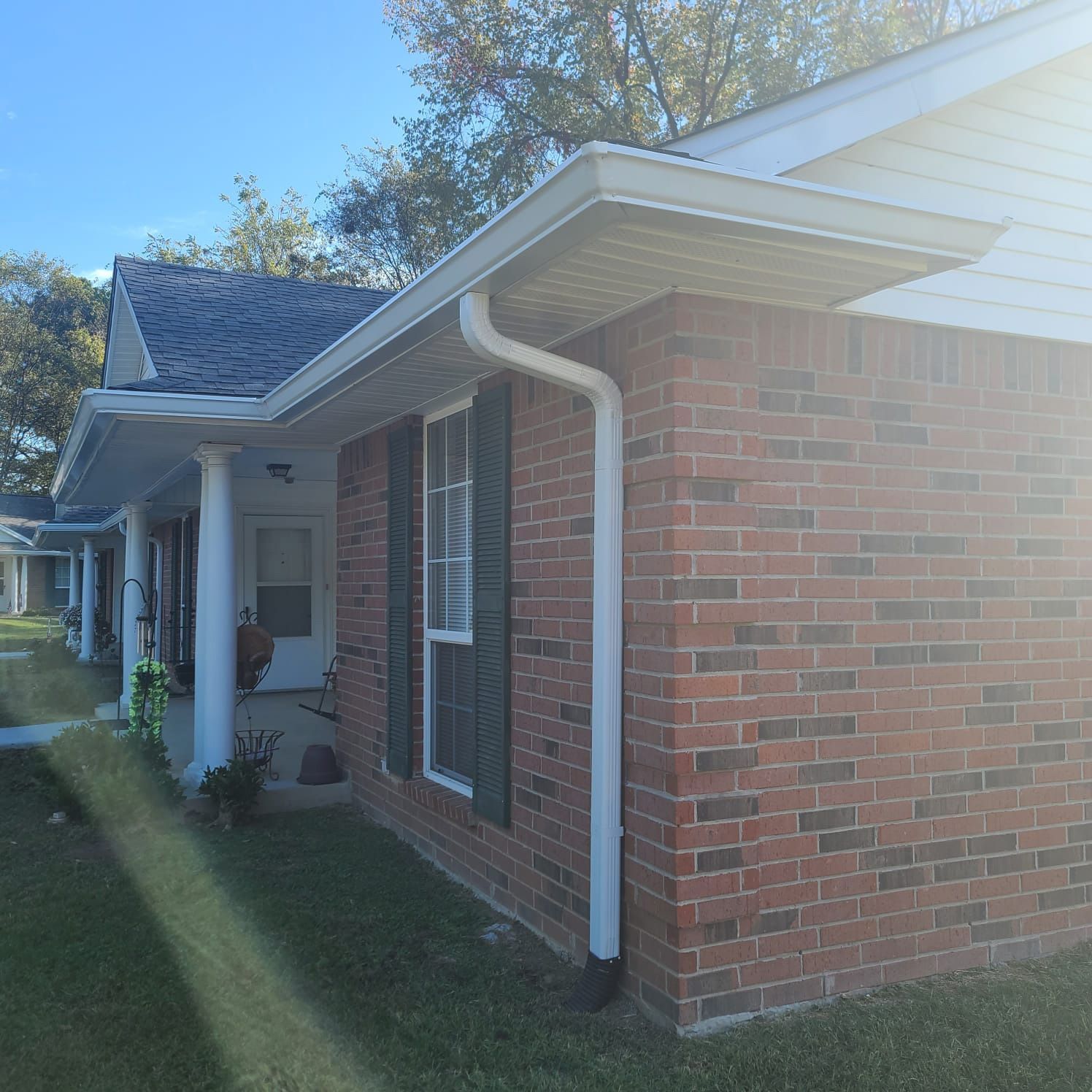 A brick residential building with a covered porch, white columns, dark shutters, and a white gutter system on a sunny day.