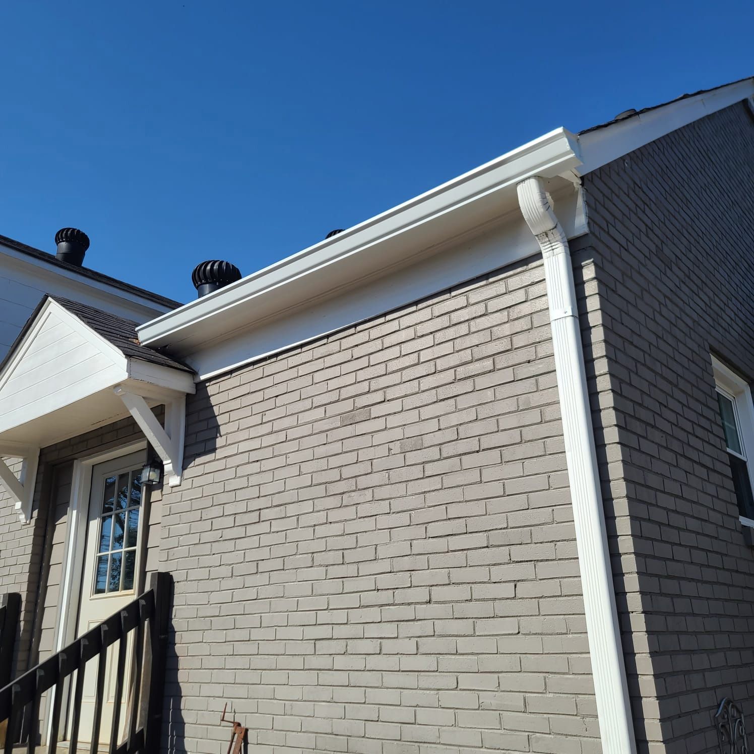 Side view of a gray brick house with white trim, a small entryway porch, and a white gutter and downspout under a blue sky.
