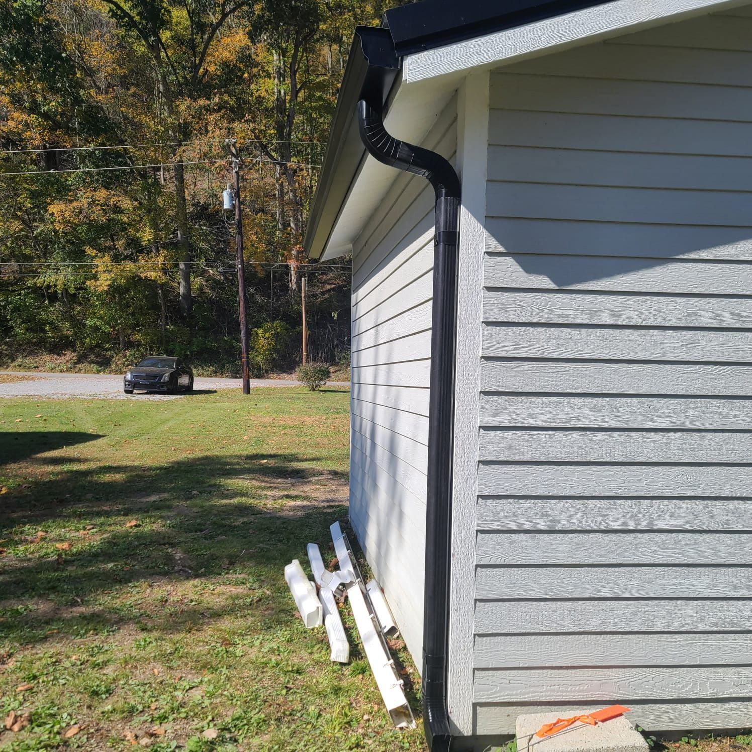 A black gutter downspout installed on the corner of a light grey house, with spare gutter pieces on the grass nearby.