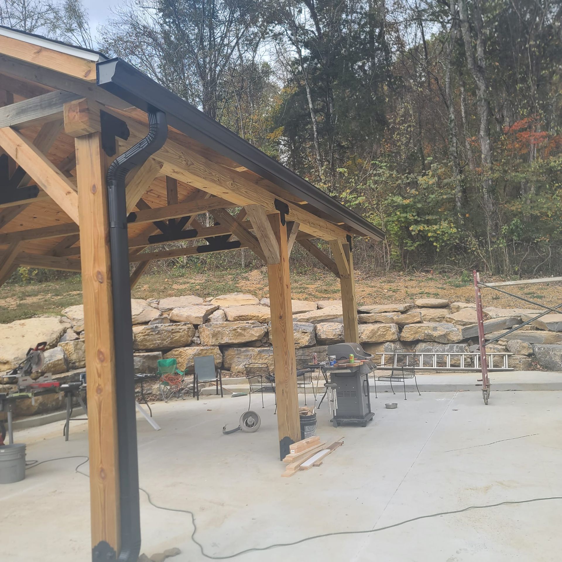 A black metal gutter and downspout installed on a timber-framed outdoor pavilion with a concrete patio.