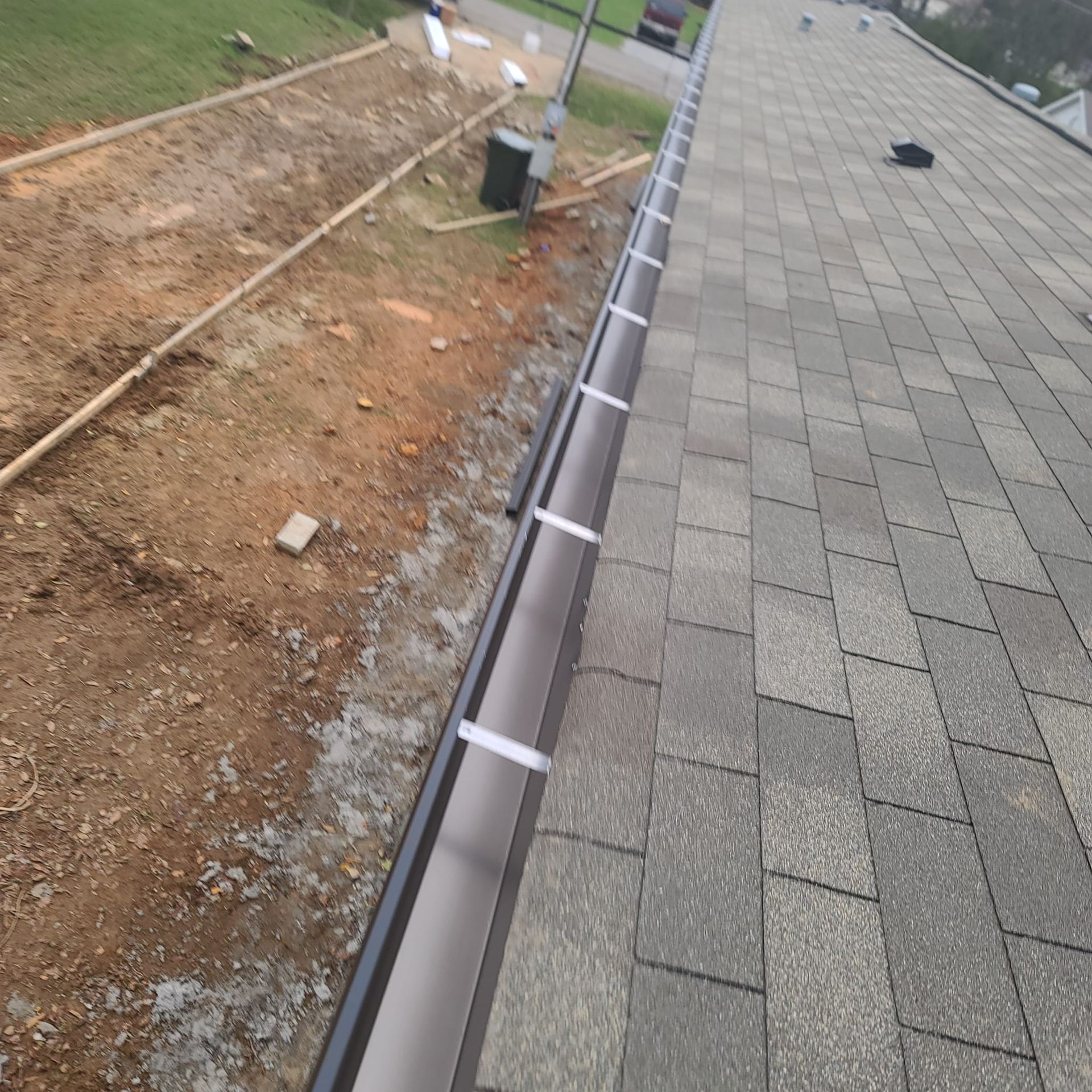A dark brown gutter installed along the edge of a grey shingled roof, adjacent to an area of exposed dirt.