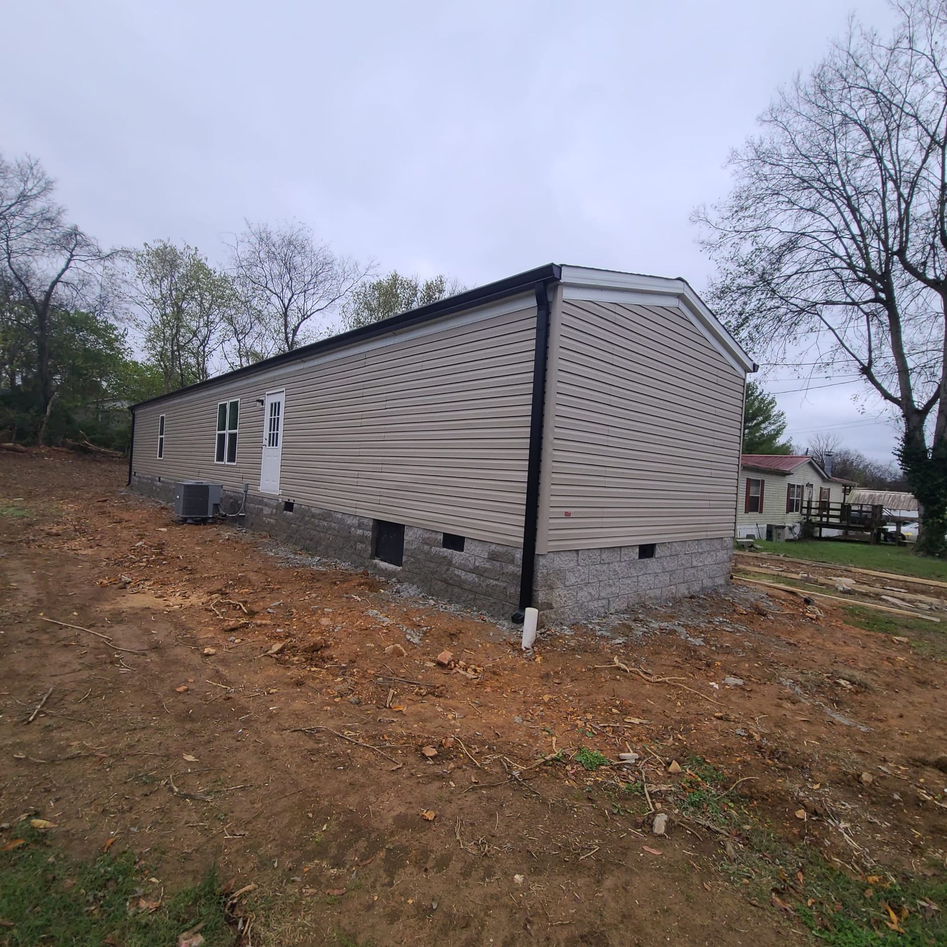 A beige mobile home with a gray stone foundation sits on a recently cleared dirt lot under an overcast sky.