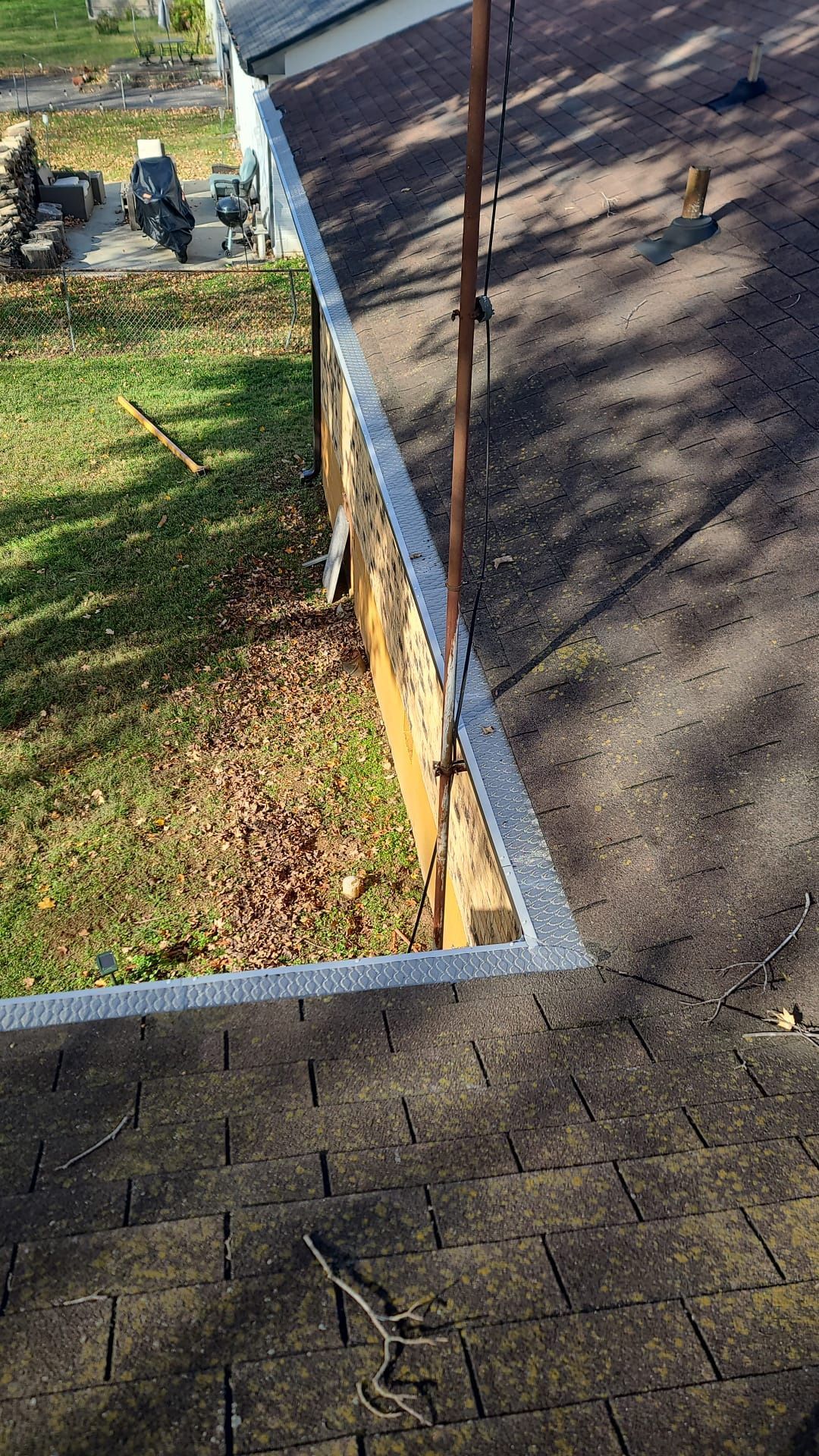 A high-angle view of a roof edge with missing shingles, exposing the underlying wooden structure and vertical piping.