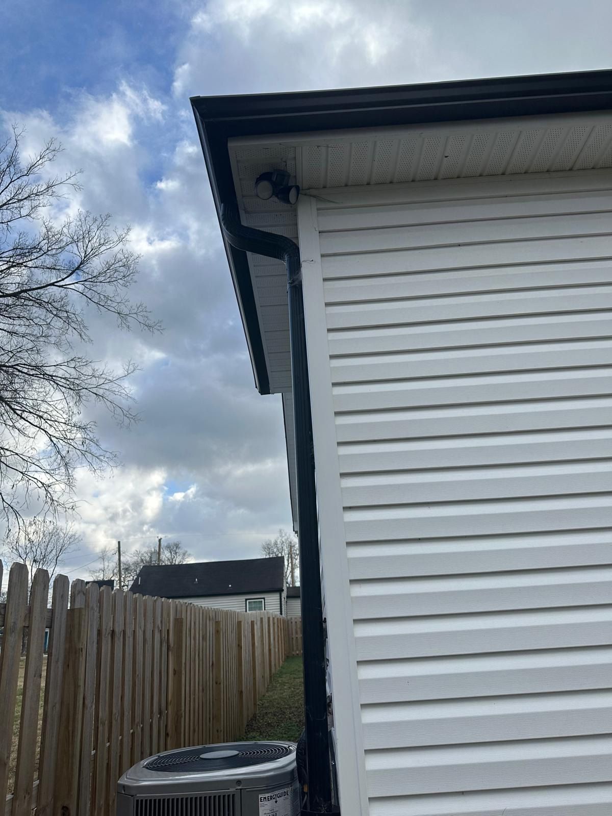 The side of a white-sided house with a black gutter corner, a wooden fence, and an AC unit under a cloudy sky.