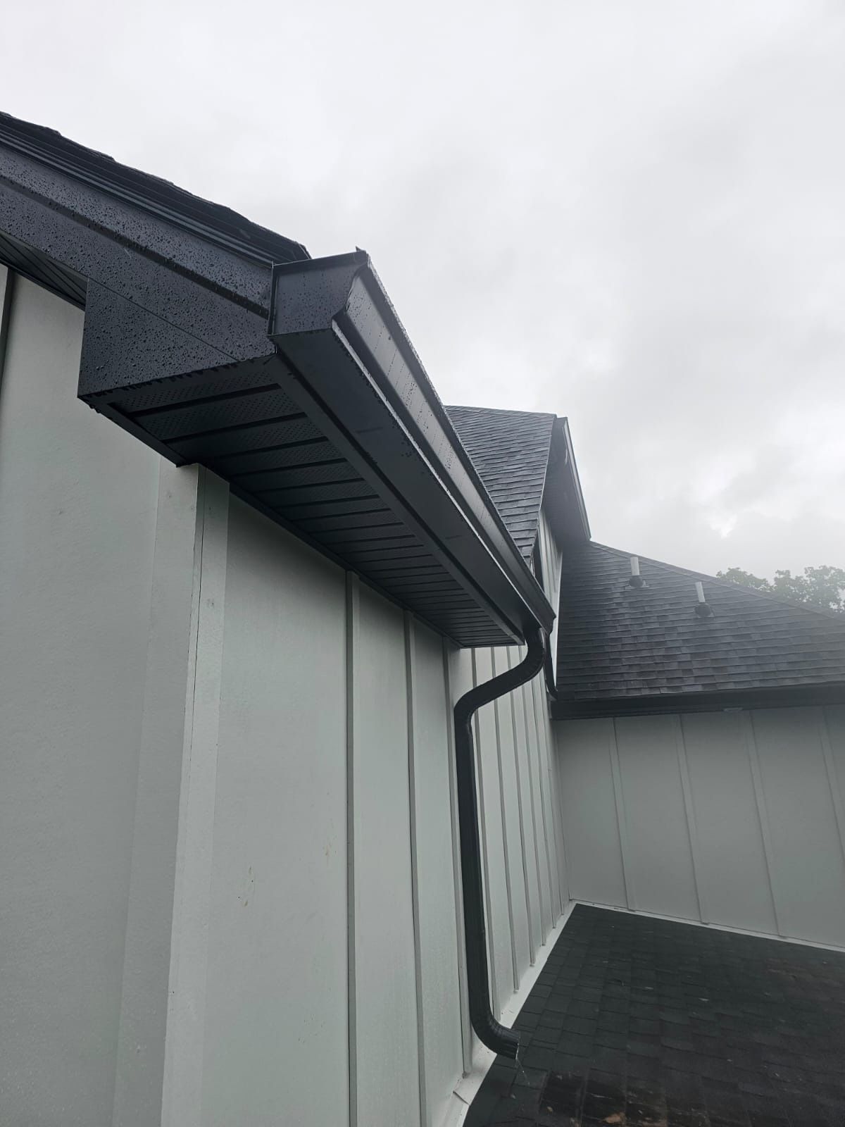 A low-angle view of a grey house exterior featuring black gutters, a black downspout, and dark shingled roofing.