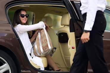 Woman in sunglasses sits in car, holding a bag, as person in suit opens the door.