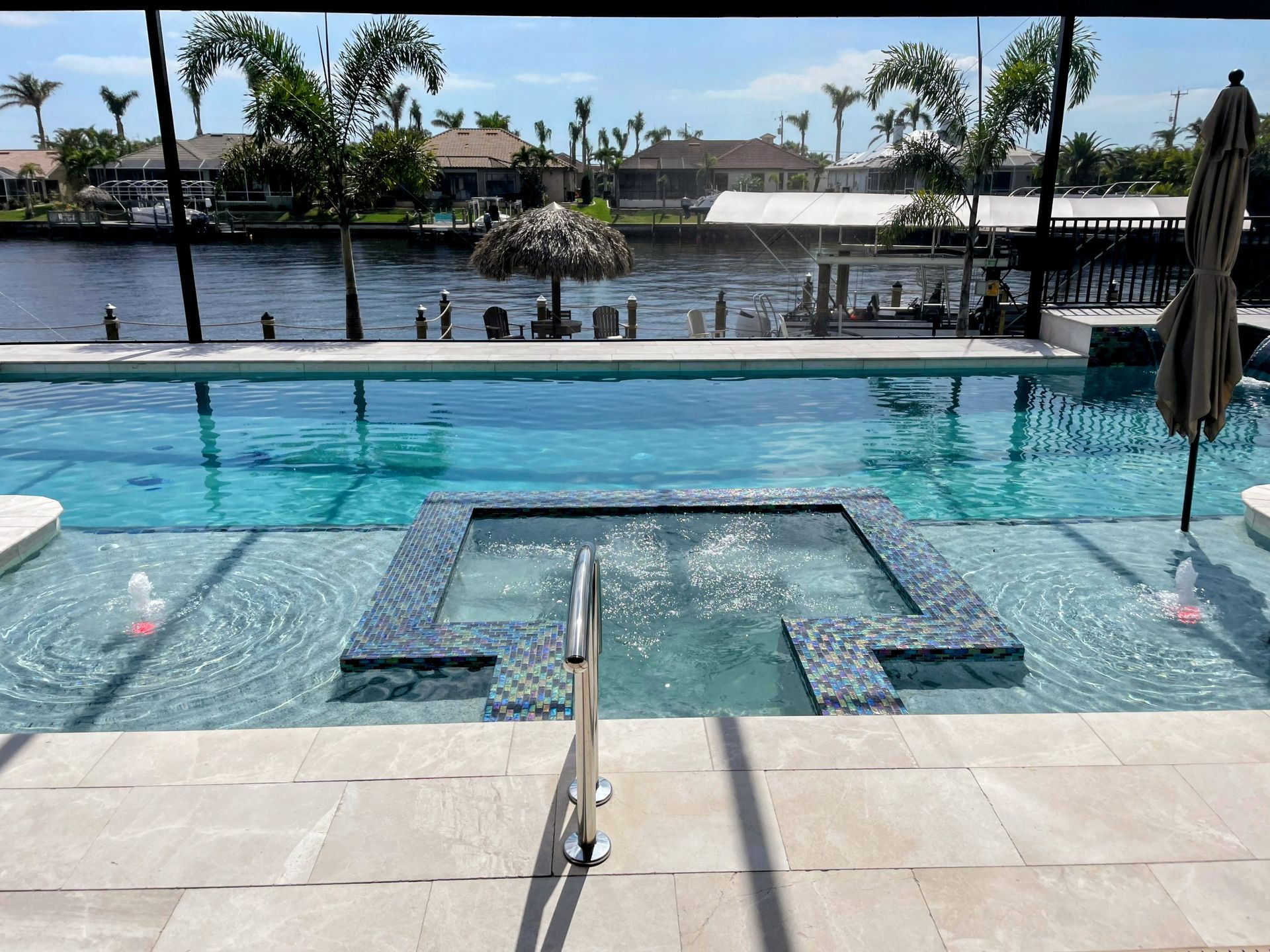 Pool with hot tub and lounge chairs overlooking a waterfront, with palm trees and umbrellas in the background.