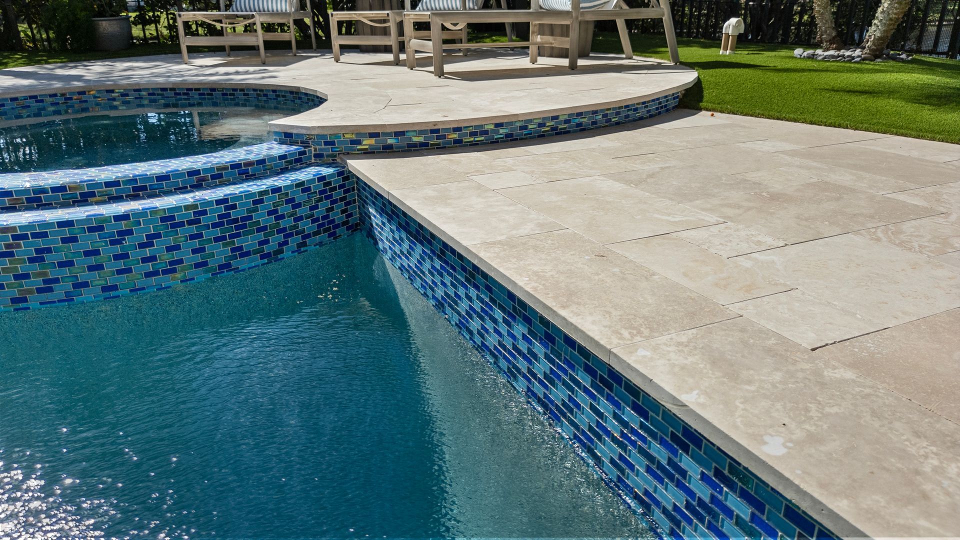 Blue-tiled swimming pool edge with concrete deck and patio seating in the background