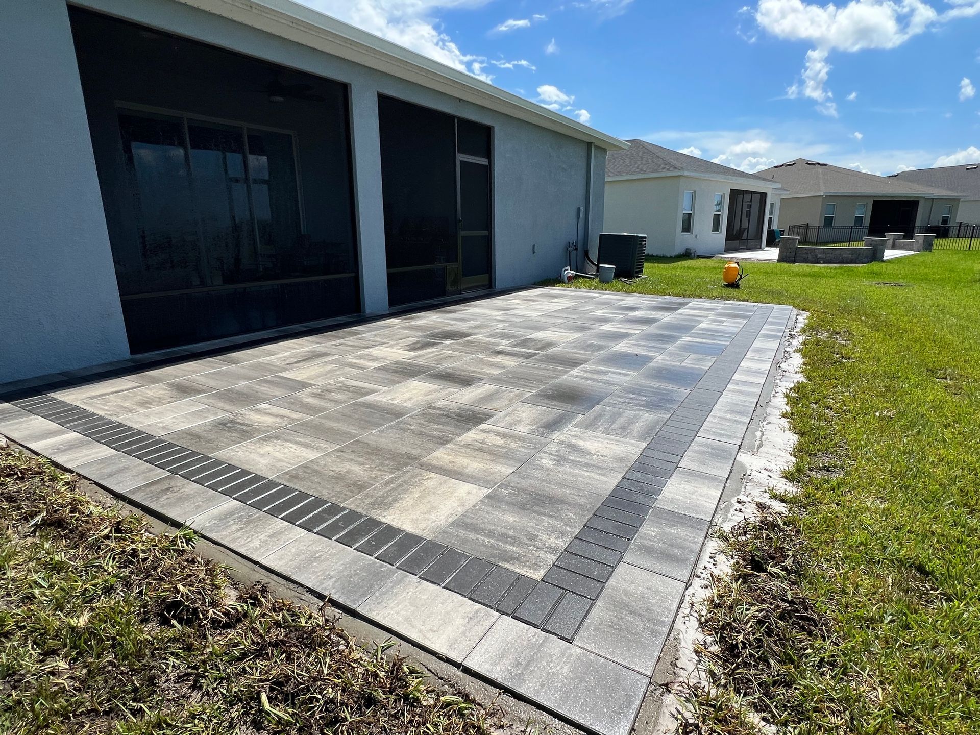 Modern tiled patio beside a house, bordered by grass and a screened porch under a bright blue sky