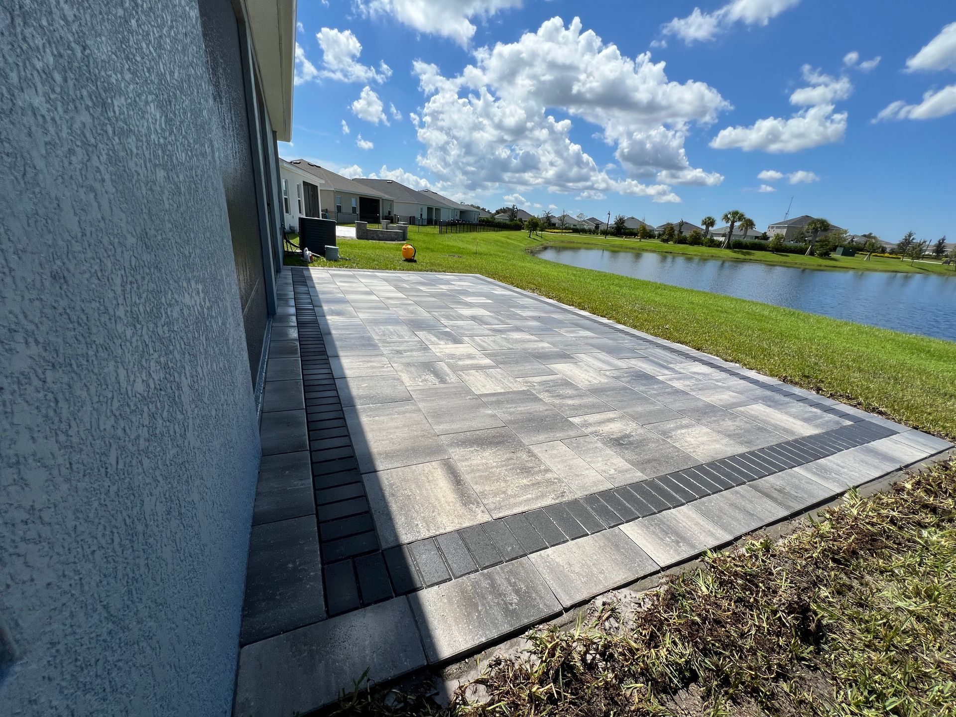 Sunlit paved patio beside a grassy pond, with a blue sky and clouds overhead.