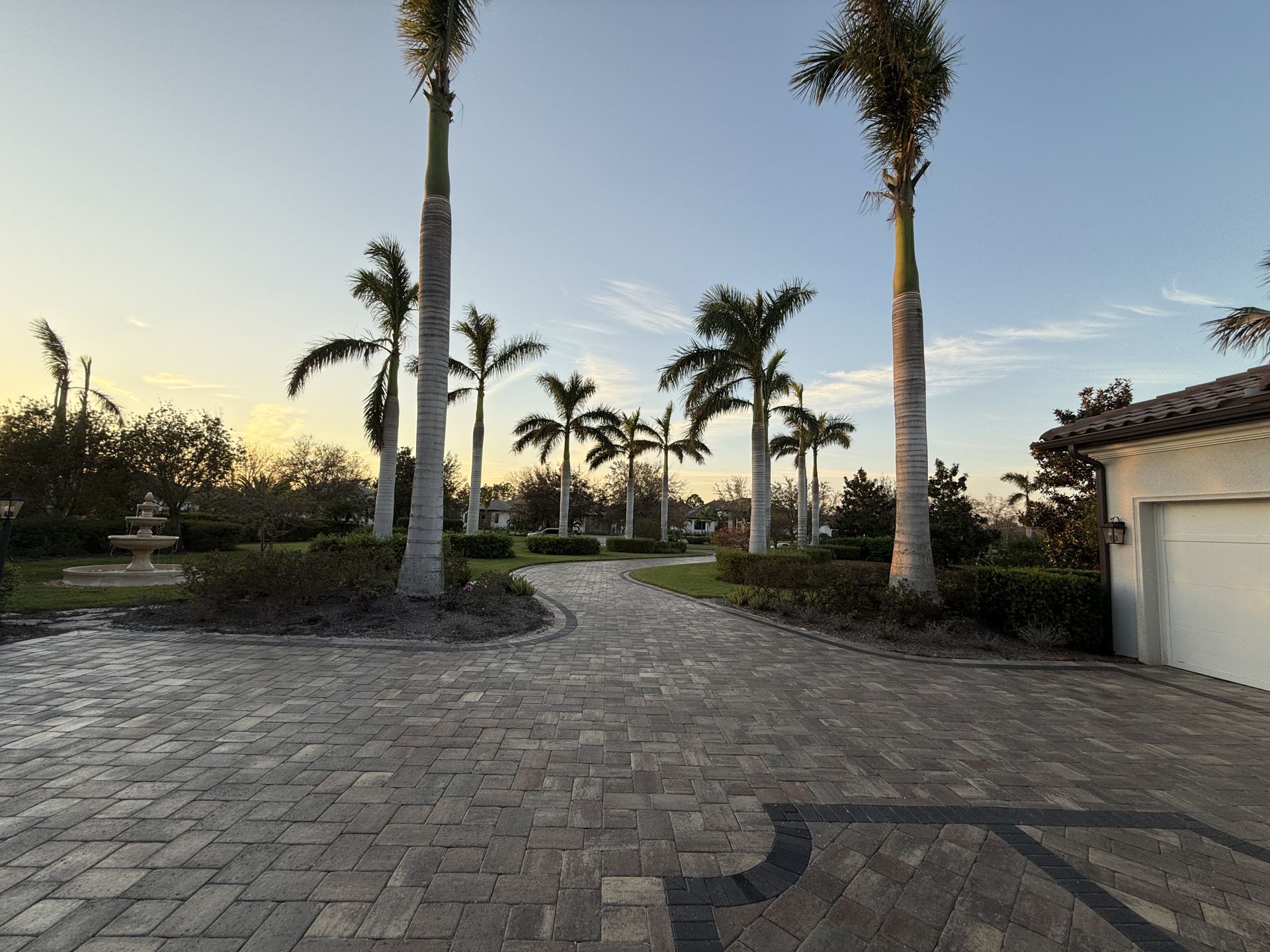 Palm-lined driveway at sunset beside a house and garage, with paved stone courtyard and landscaped garden.