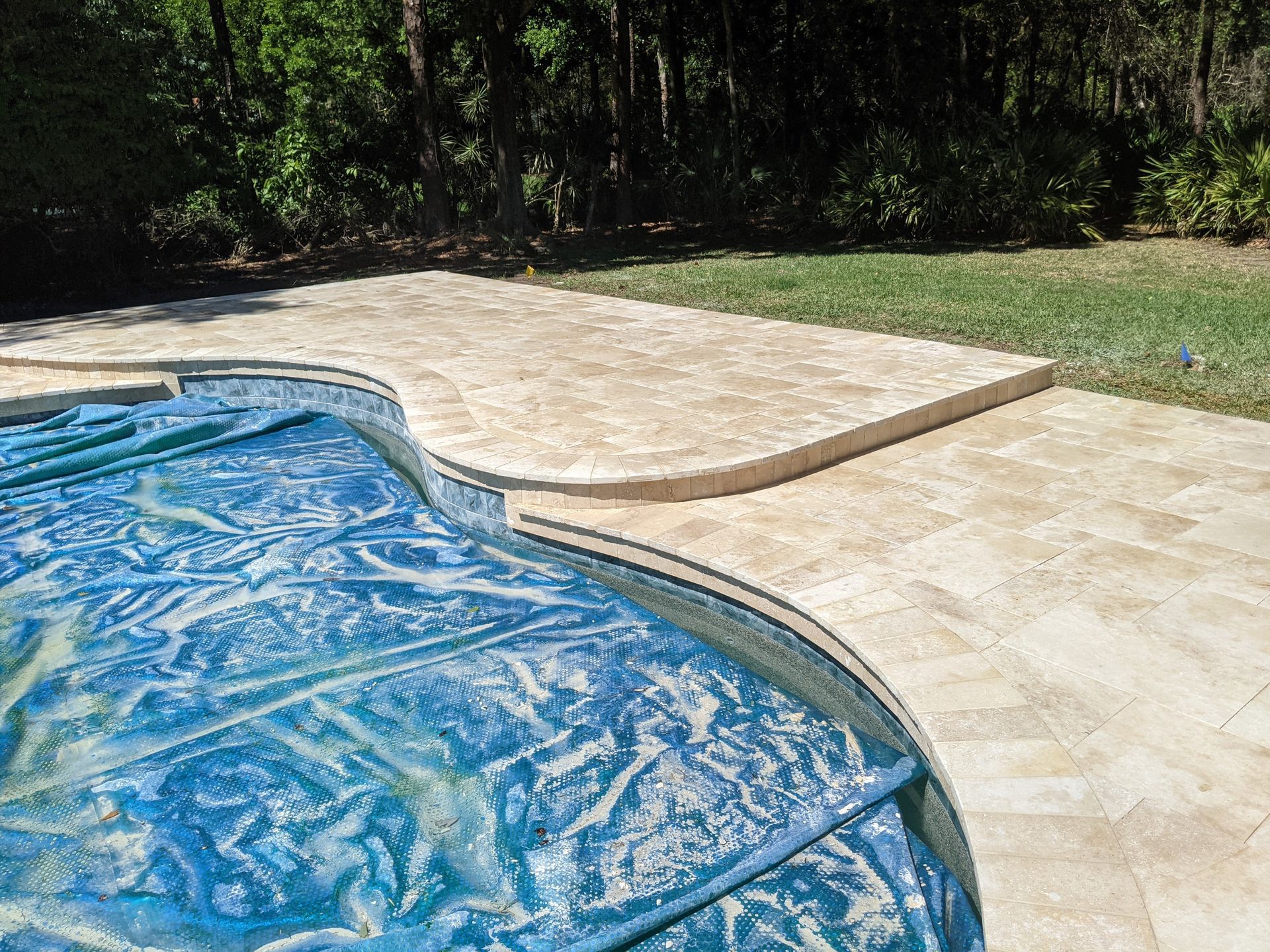 Poolside patio with tan stone decking beside a blue swimming pool and grassy yard.