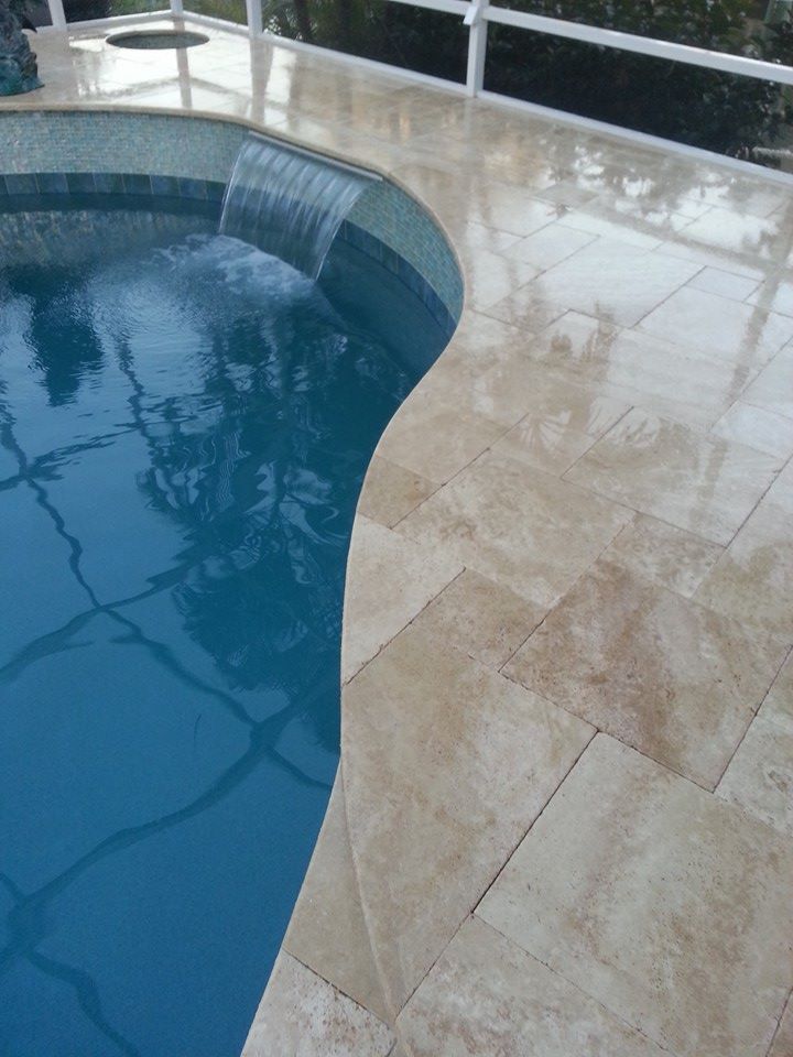 Curved blue swimming pool beside a beige stone deck with water reflections
