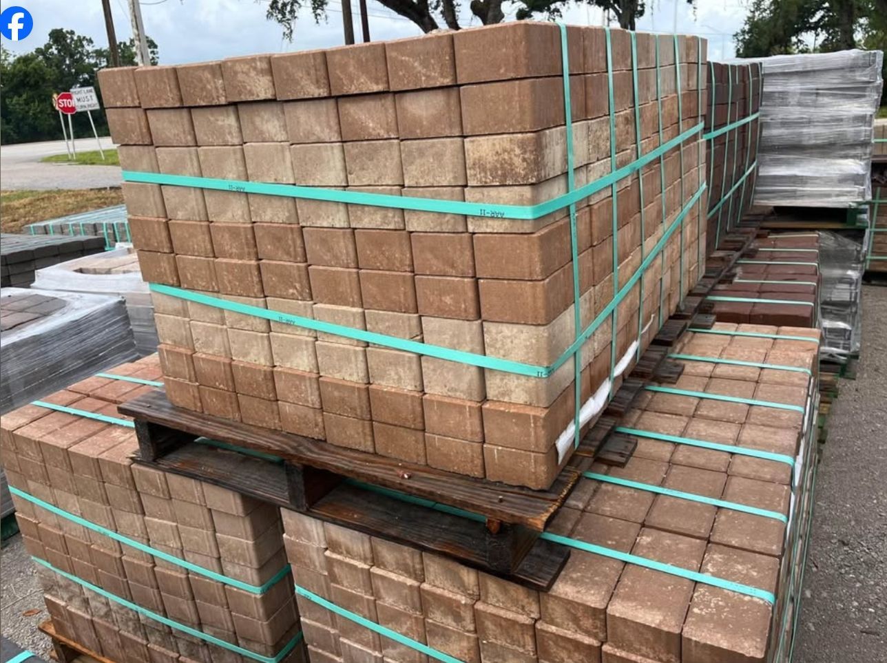 Stacked bricks bound with green straps on pallets outdoors, with wrapped materials in the background