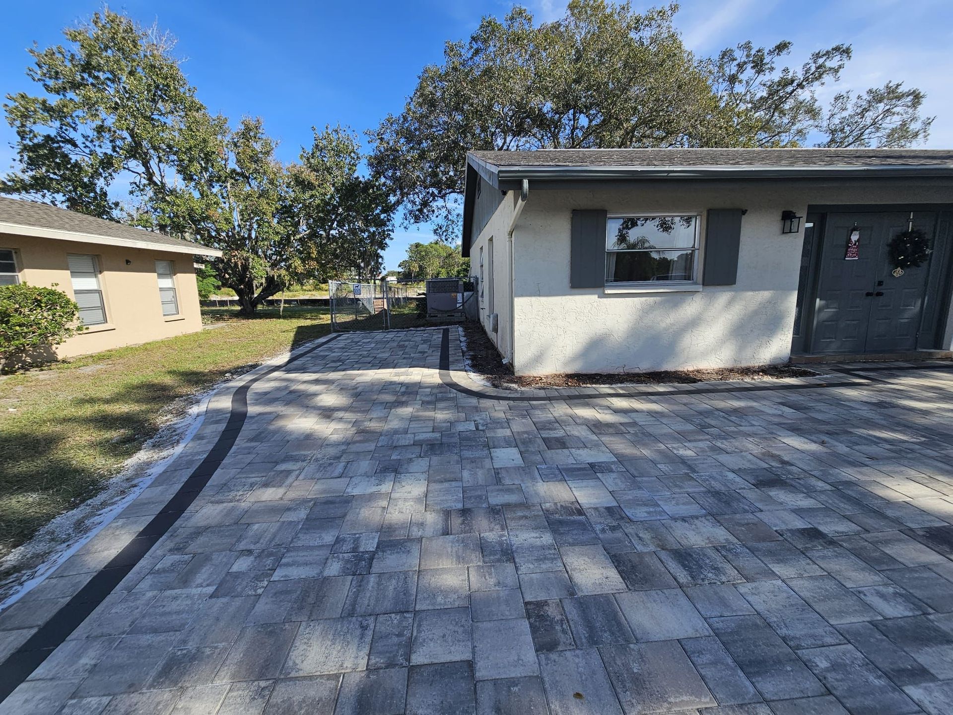 Paver driveway and courtyard beside a small house, with trees and a gate in the background.