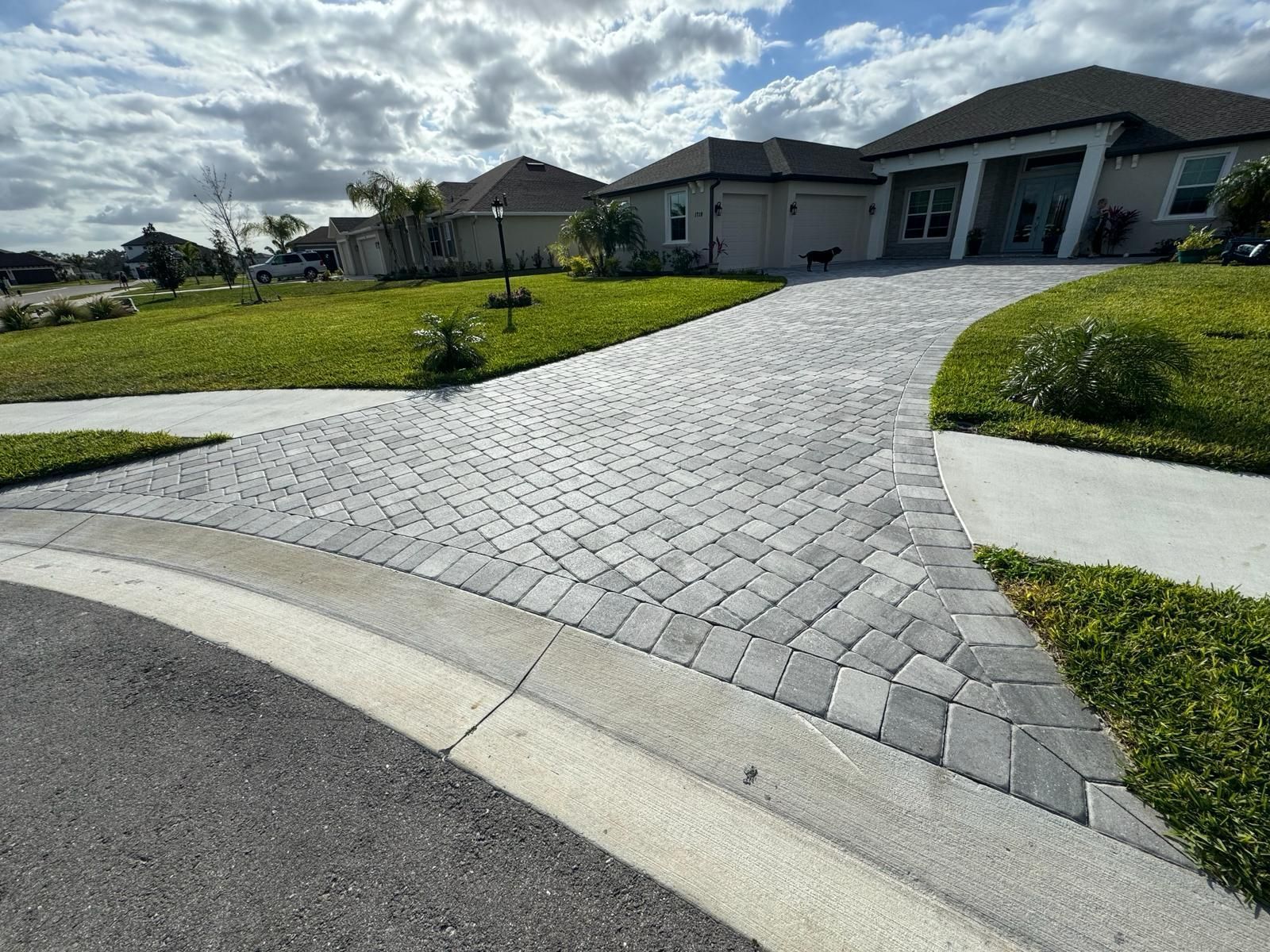 Curved gray paver driveway leading to a modern house with green lawn and cloudy sky.