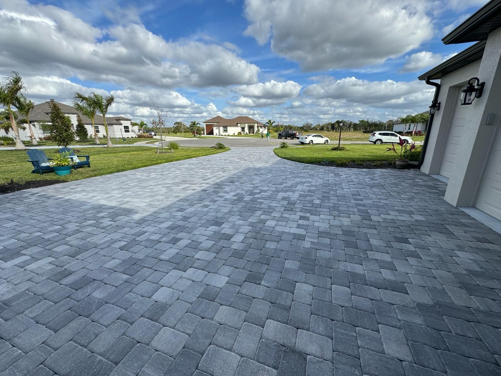 Paved suburban driveway and patio under cloudy sky, with houses and lawn in the background.