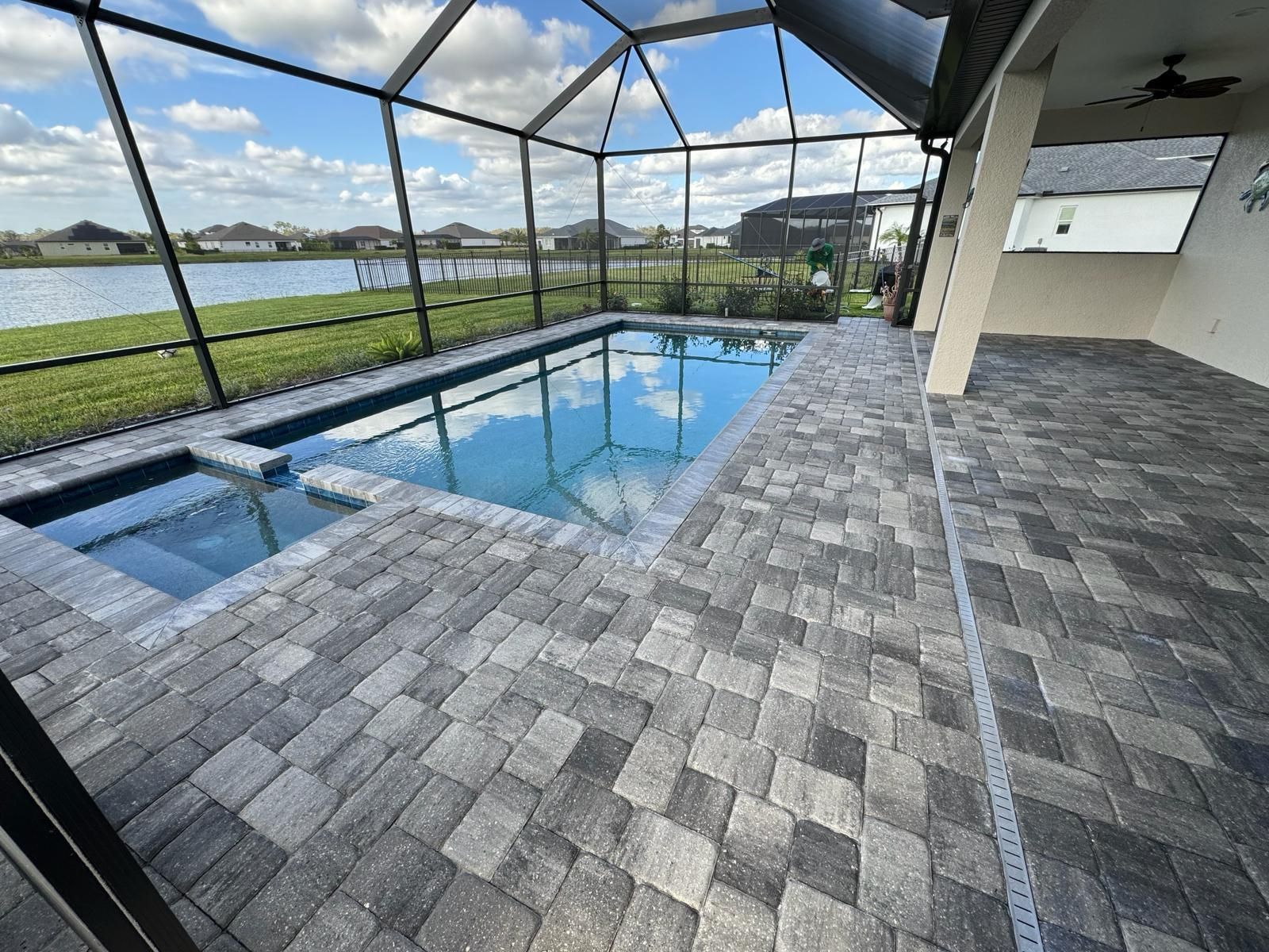 Covered outdoor pool beside a gray paver patio, enclosed by a glass screen and overlooking a grassy field.