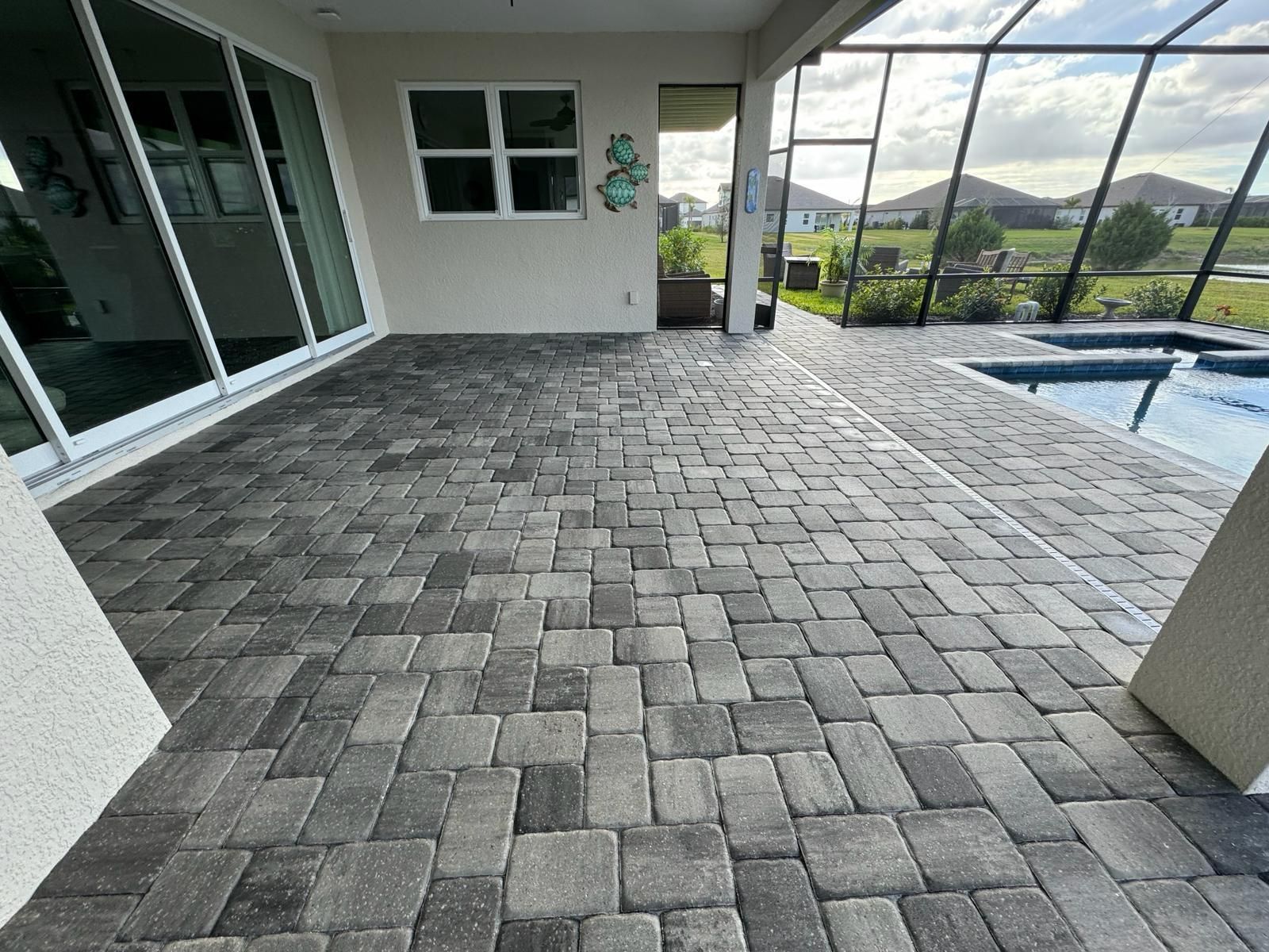 Covered patio with gray paver flooring, sliding glass doors, and a pool overlooking a grassy landscape.