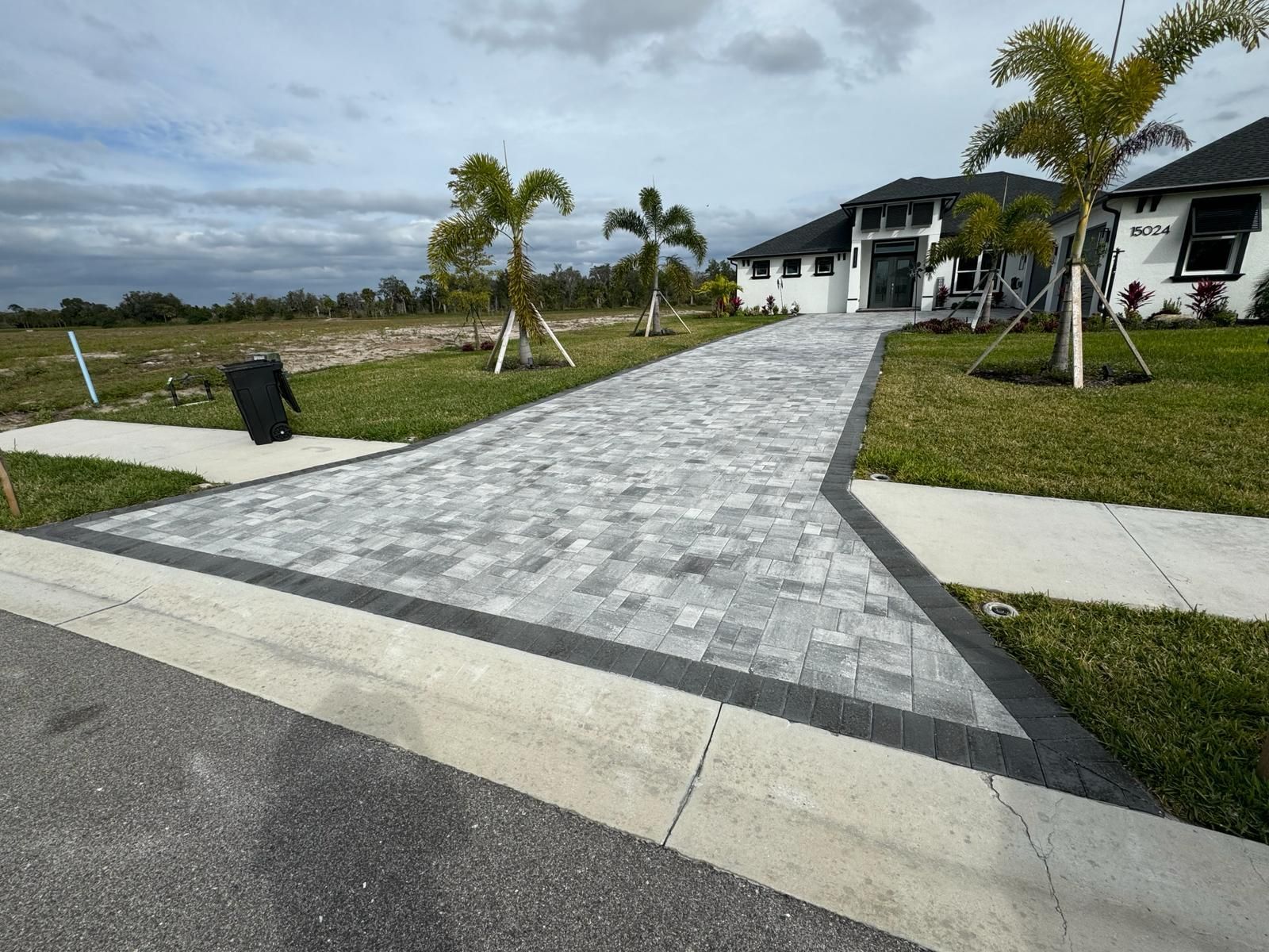 Stone walkway leads to a white building beside palm trees and grassy landscaping under a cloudy sky.