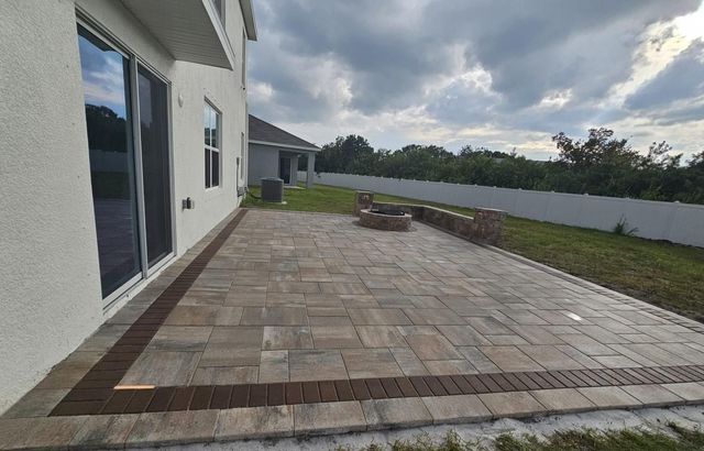 Wide stone patio beside a white house, with a grass border and cloudy sky in the background.