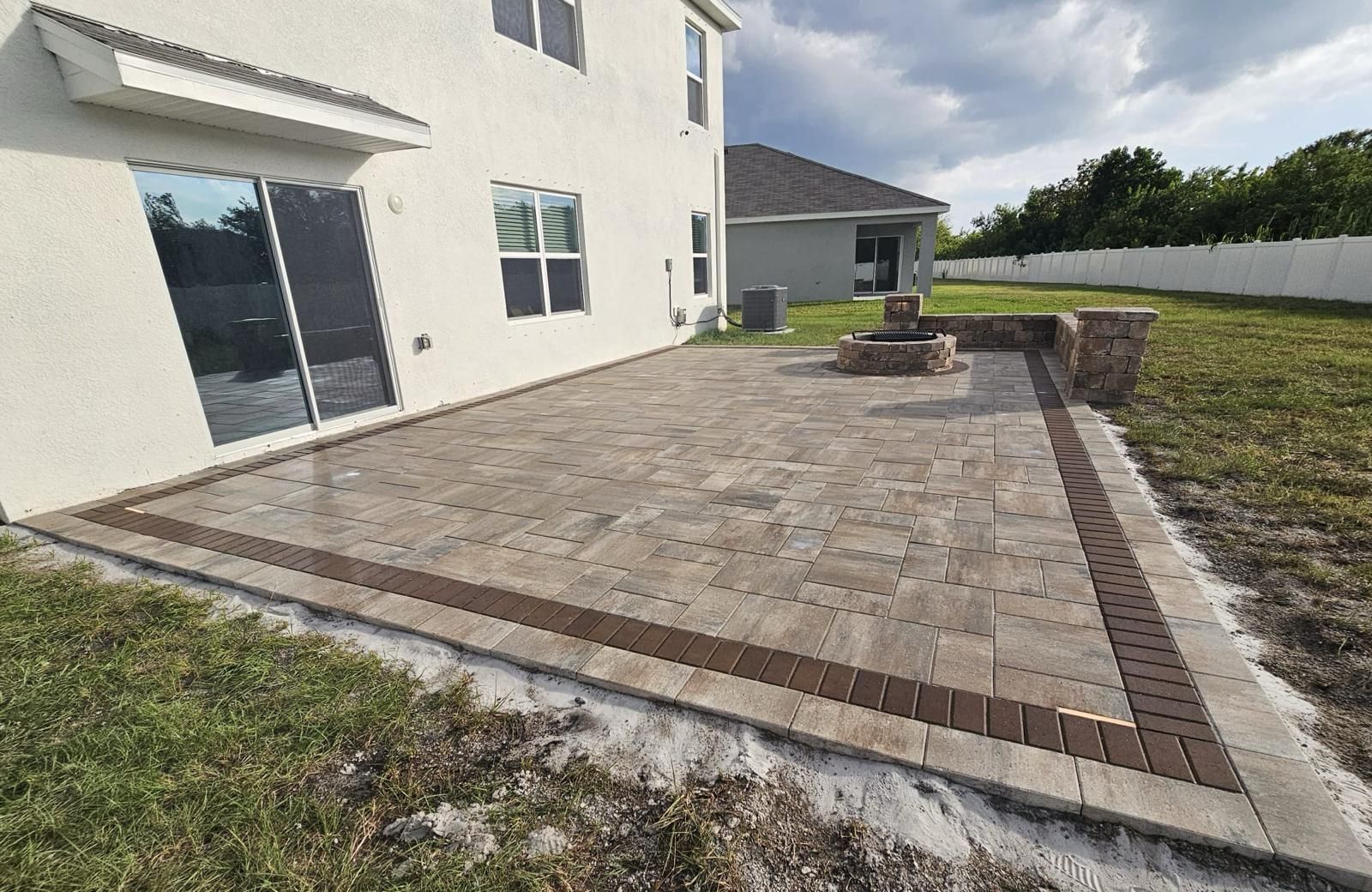 Paver patio beside a white house with a central fire pit and landscaped border