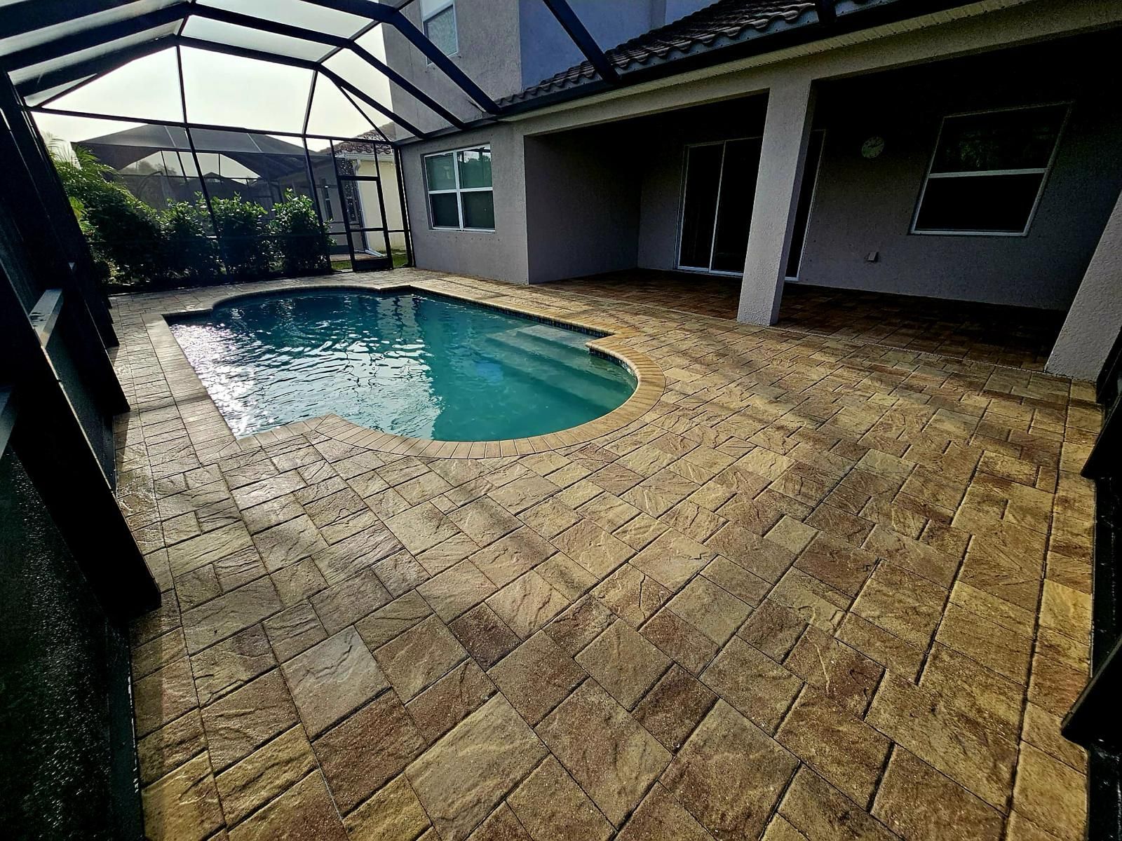 Covered patio with a small turquoise swimming pool and tan paver deck beside a house