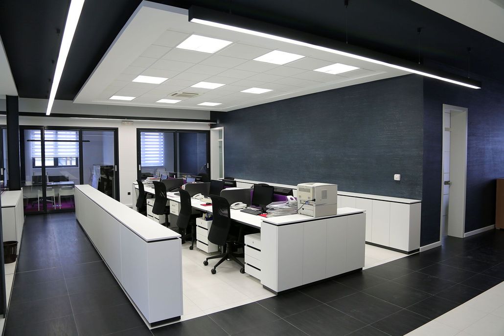 Modern office with desks, computers, black chairs, and a gray accent wall beneath bright ceiling lights